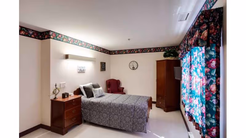 A neatly arranged bedroom in a senior living facility with a single bed covered in a gray patterned bedspread, a wooden nightstand with a lamp and decorative items, a red upholstered armchair in the corner, a wooden wardrobe, a wall clock, and floral curtains on the window.