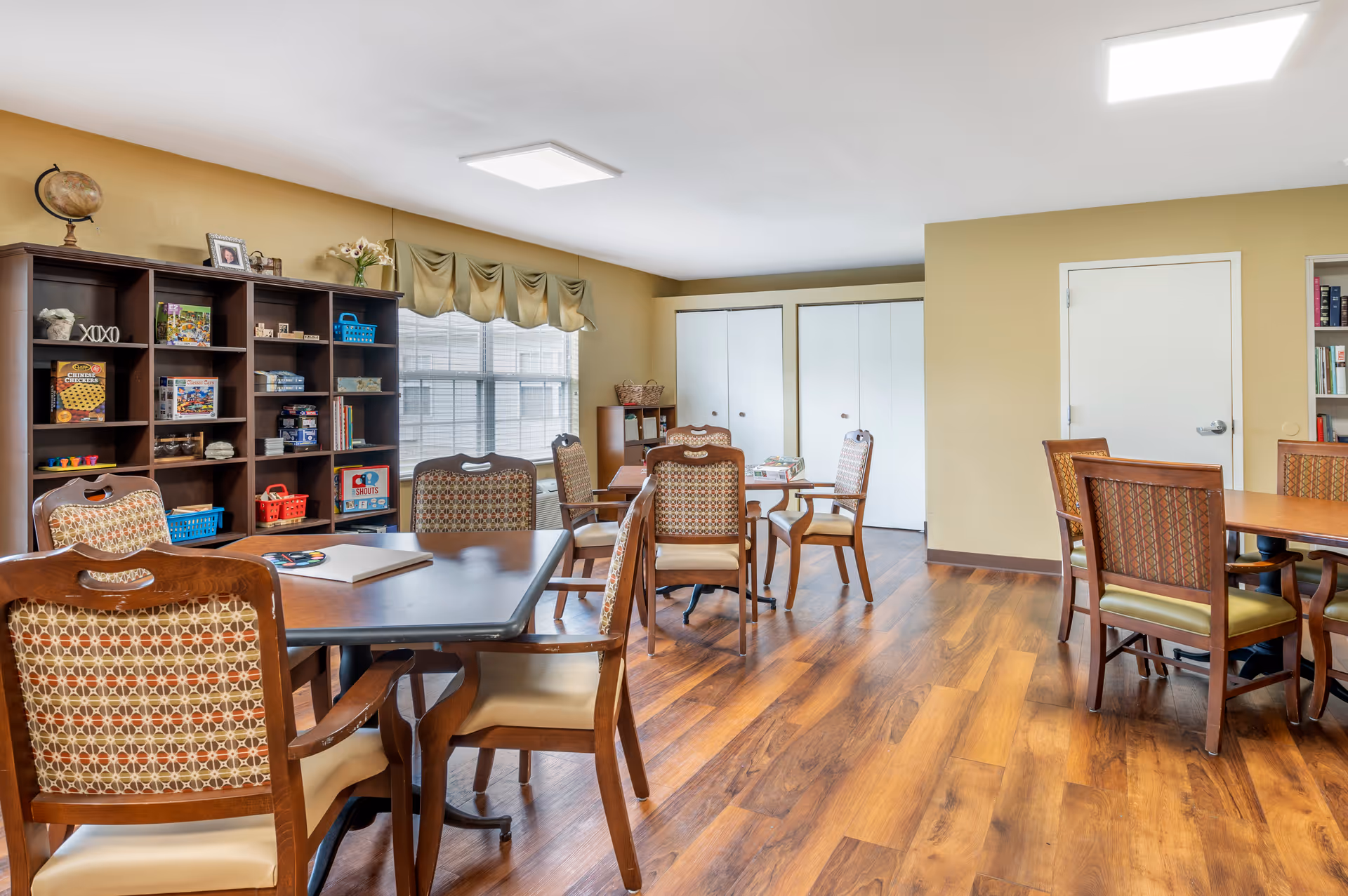 A bright room with wooden flooring and beige walls, furnished with several wooden tables and chairs with patterned upholstery. A large window with beige curtains lets in natural light. There is a dark wooden shelving unit filled with board games and decorative items, and white double-door closets along the back wall. The room appears to be a communal activity or game room.