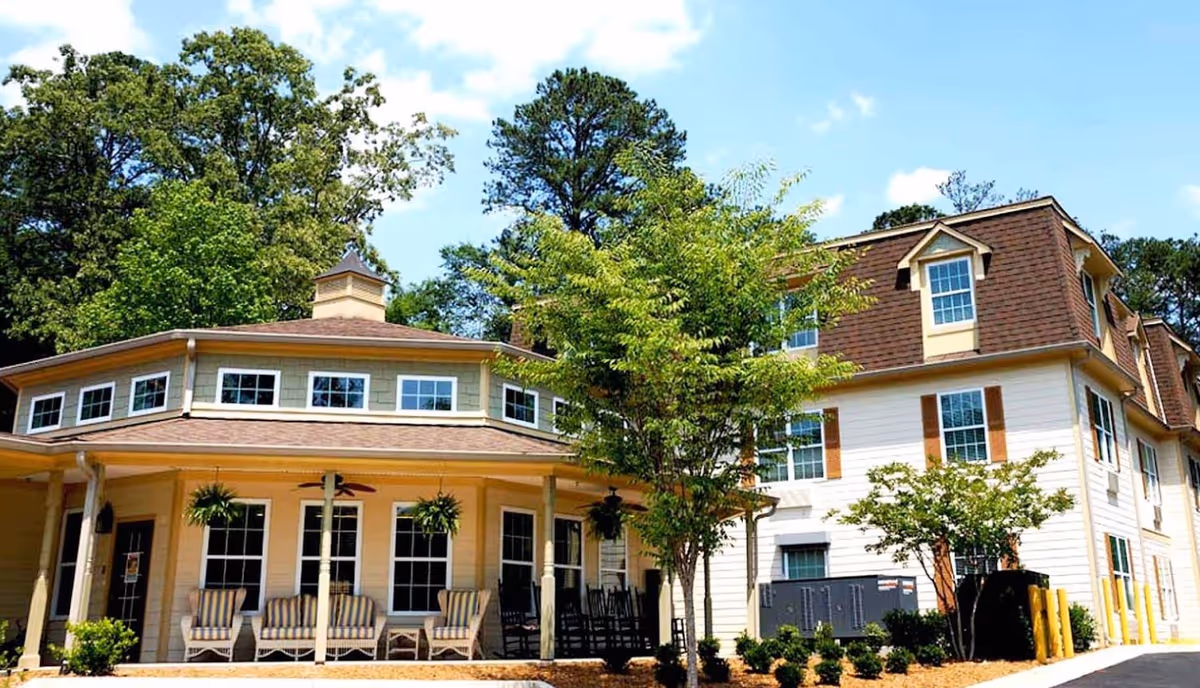 Exterior view of a senior living facility building with a covered porch featuring several chairs and hanging plants. The building has multiple windows, a brown shingled roof, and is surrounded by trees and landscaping under a blue sky with some clouds.