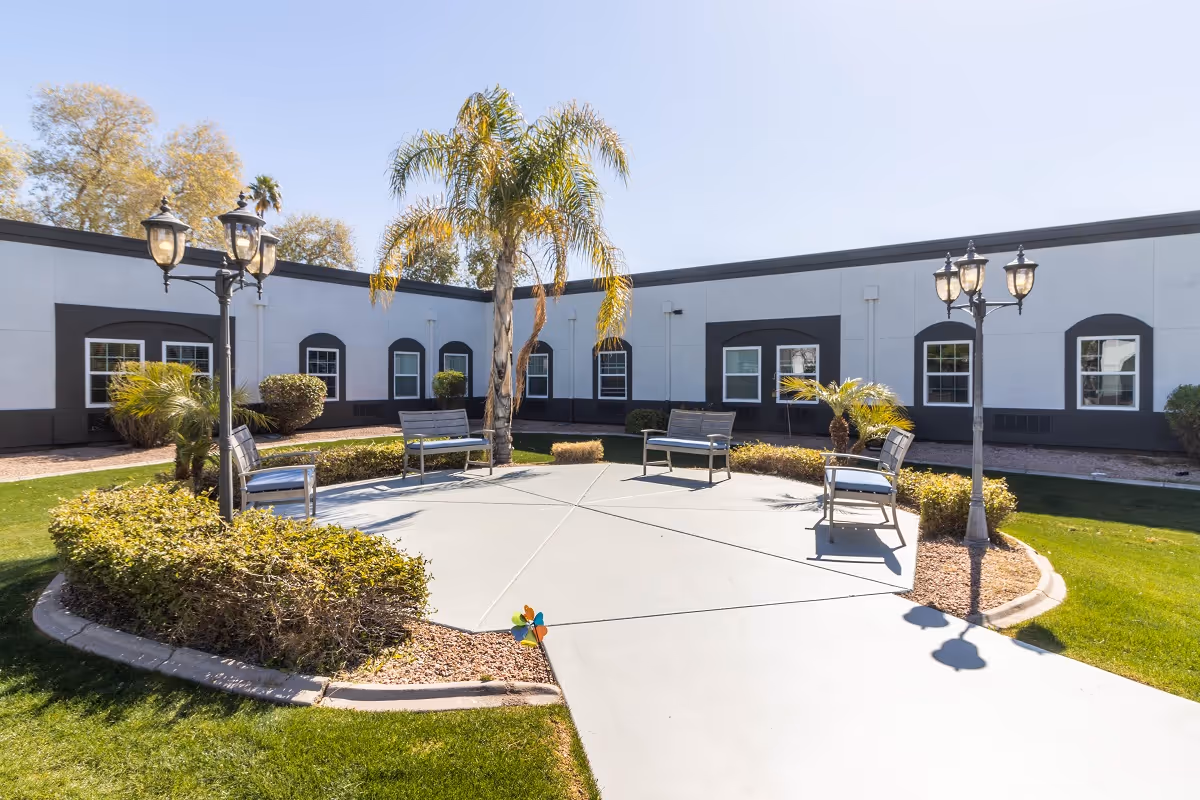 Outdoor courtyard area with a circular concrete seating space surrounded by bushes and palm trees. Four metal benches with cushions are arranged around the circle. Two vintage-style street lamps are positioned on opposite sides of the seating area. The courtyard is enclosed by a single-story building with multiple windows.