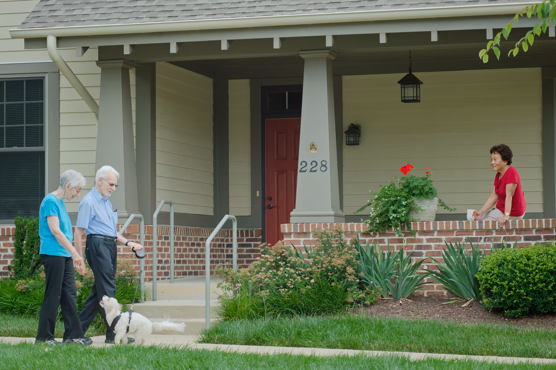 Two elderly people walking a small white dog on a leash on a sidewalk in front of a house with a red door and the number 228. Another elderly woman is sitting on the brick porch wall, smiling and watching them. The house has beige siding, a covered porch, and some green plants and flowers in front.