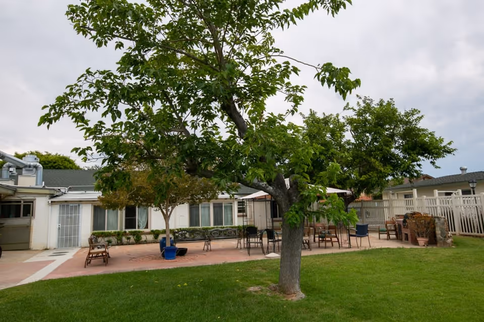 Outdoor patio area at Lo-Har Senior Living featuring green grass, several trees, patio furniture including tables and chairs, and a brick barbecue grill, all surrounded by a white fence and adjacent to a single-story building.
