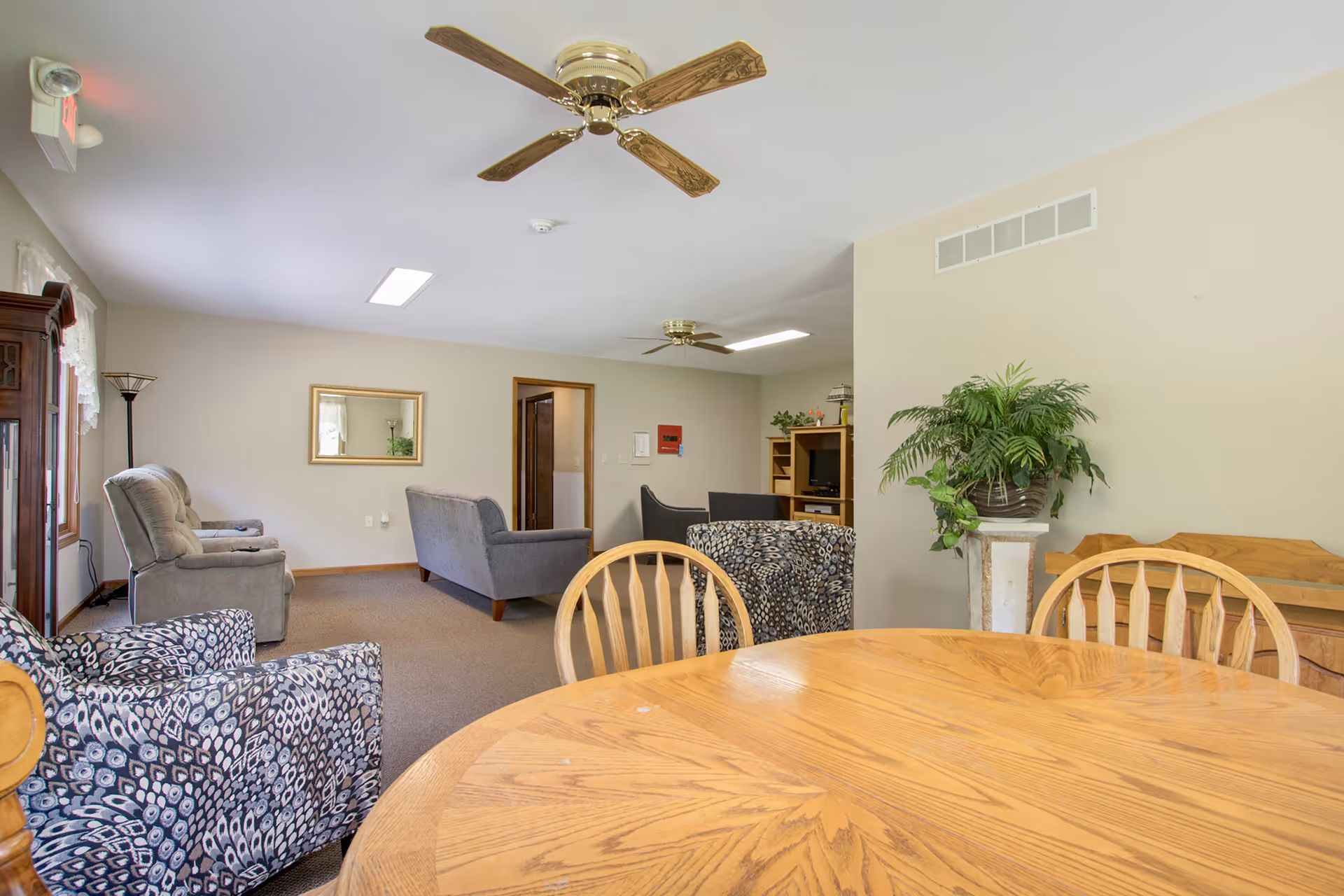 A spacious living room area in Pine Ridge Assisted Living featuring a round wooden table with chairs in the foreground, patterned armchairs, gray upholstered sofas, a wooden entertainment center with a TV, a tall plant on a pedestal, and ceiling fans. The room has beige walls, carpeted floors, and a window with lace curtains allowing natural light.