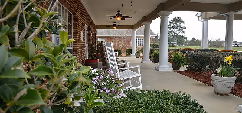 Covered front porch with white rocking chairs, columns, potted plants, and landscaped shrubs at a nursing facility.