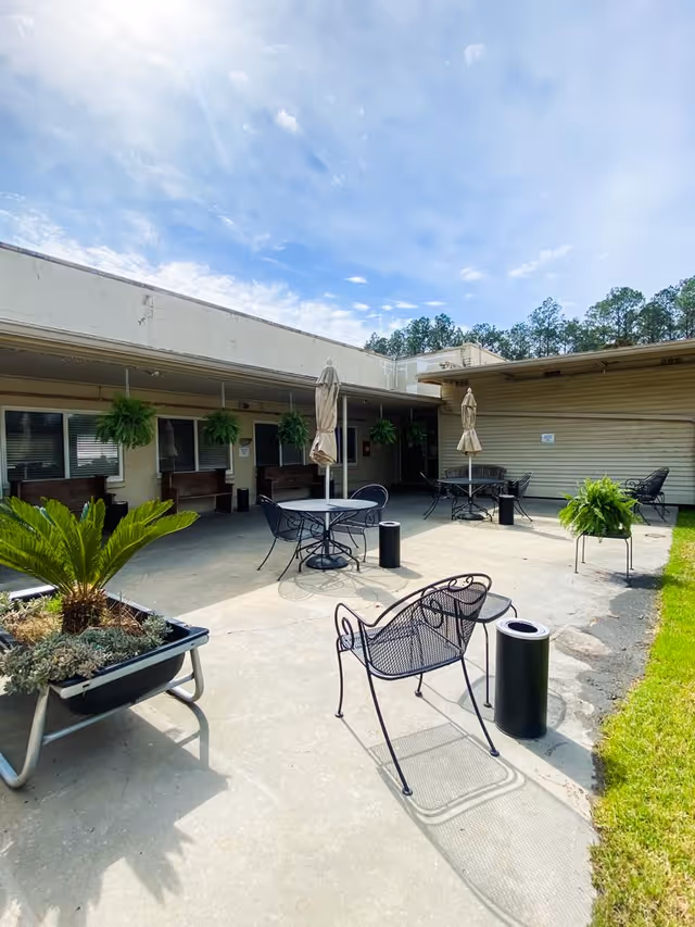 Outdoor patio area at Picayune Rehabilitation and Healthcare Center with metal chairs and tables, some with closed umbrellas. Several hanging plants and potted greenery are visible. The patio is adjacent to a building with windows and benches under an overhang. The sky is partly cloudy.