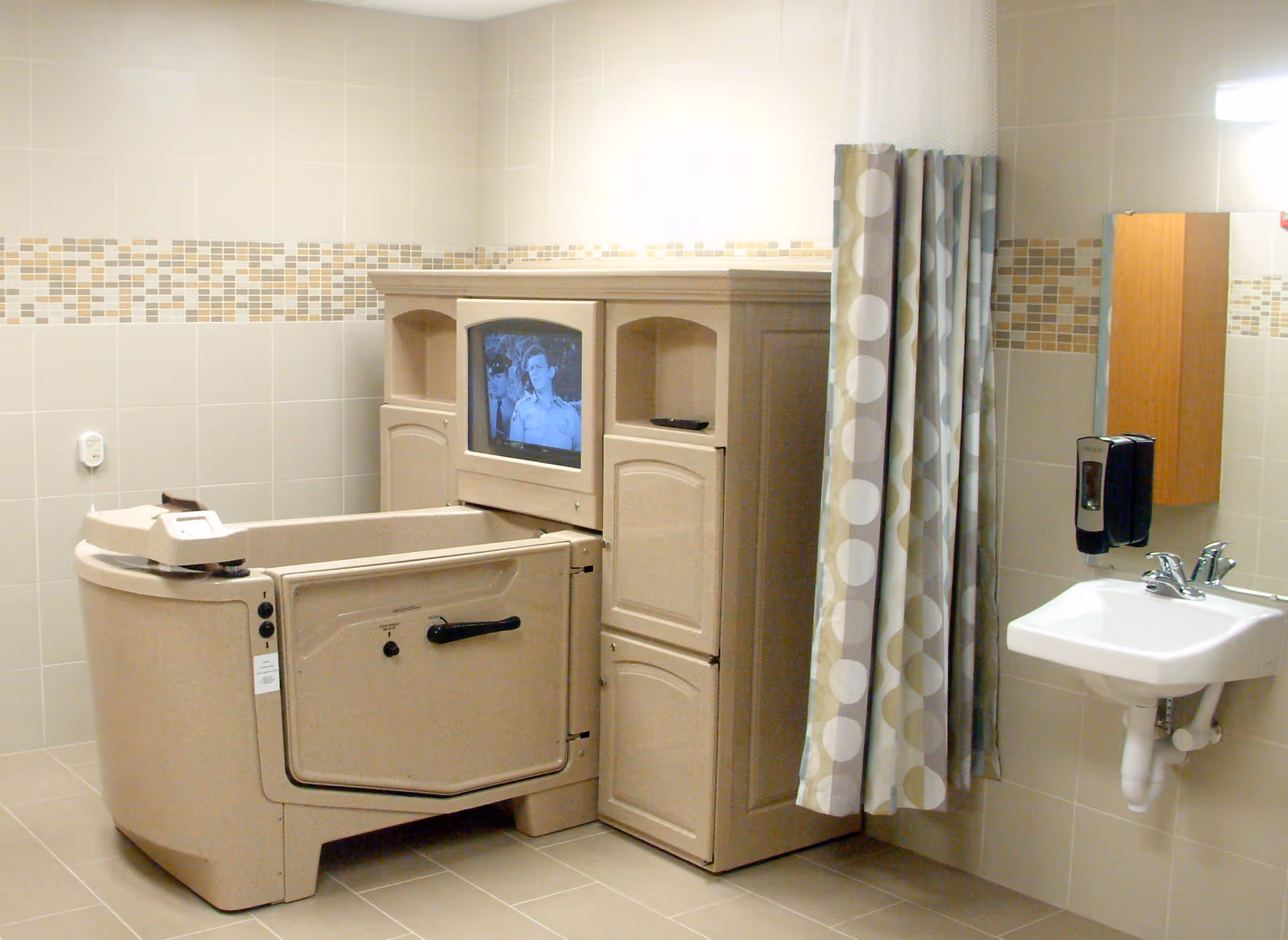 A specialized bathing room in a senior care facility featuring a beige walk-in bathtub with a door, a built-in television screen, a patterned privacy curtain, a wall-mounted soap dispenser, a white sink with a faucet, and beige tiled walls with a decorative mosaic tile border.