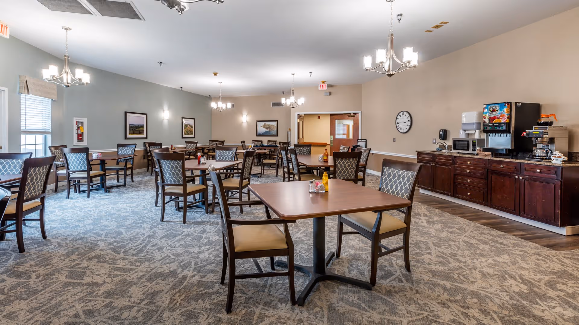 A spacious dining room with multiple square wooden tables and cushioned chairs arranged neatly. The room features soft lighting from ceiling chandeliers, framed artwork on the walls, and a carpeted floor with a subtle pattern. Along one wall, there is a dark wooden cabinet with a coffee machine, microwave, and other beverage supplies. The room has large windows with blinds and an exit sign above a door.