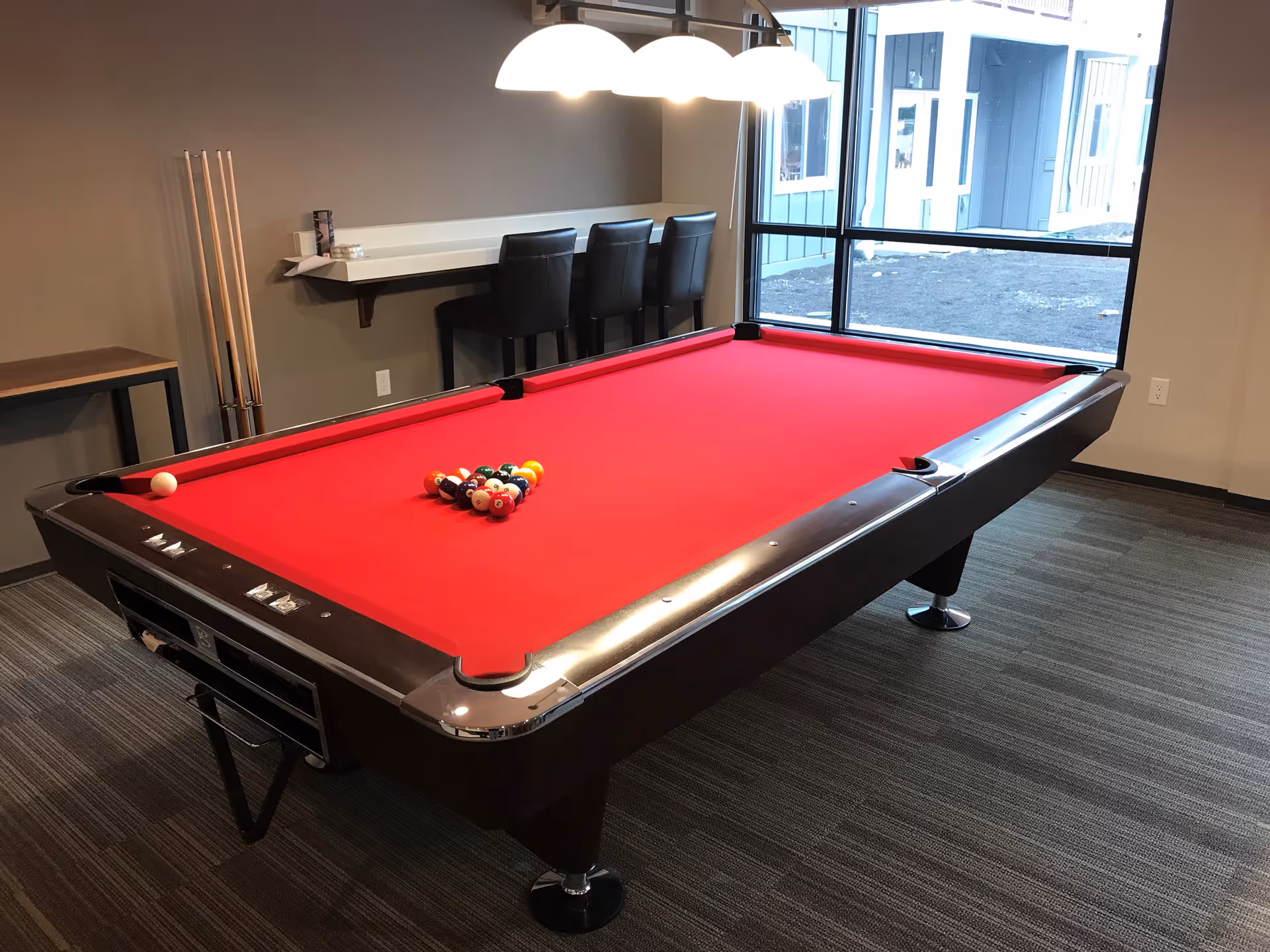 Red-felt pool table in a game room with cue sticks, a counter with stools, and a large window.