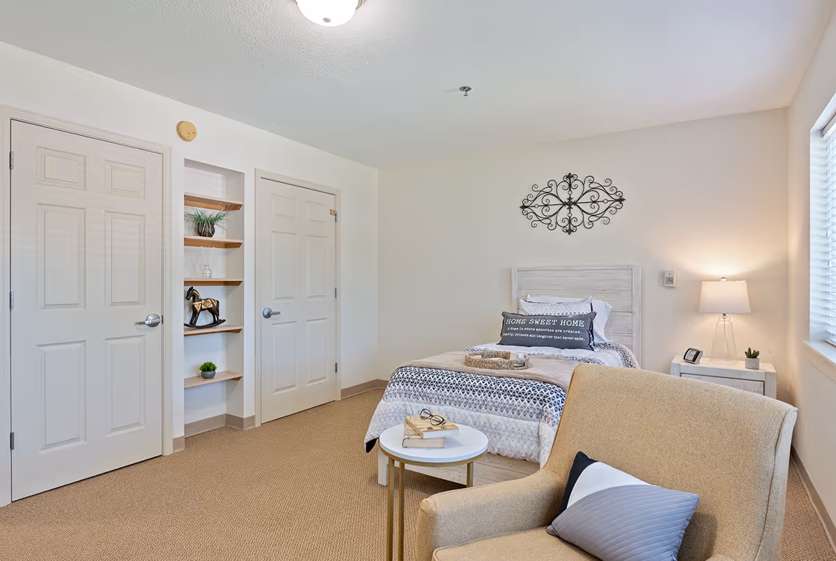 A cozy bedroom in Highlands Square featuring a single bed with patterned bedding and a decorative pillow that reads 'Home Sweet Home.' There is a beige armchair with a pillow in the foreground, a small round white table with books and glasses, a nightstand with a lamp and small plant, two closed doors, and built-in shelves with decorative items. The room has beige carpeting and white walls with a decorative metal wall art above the bed.
