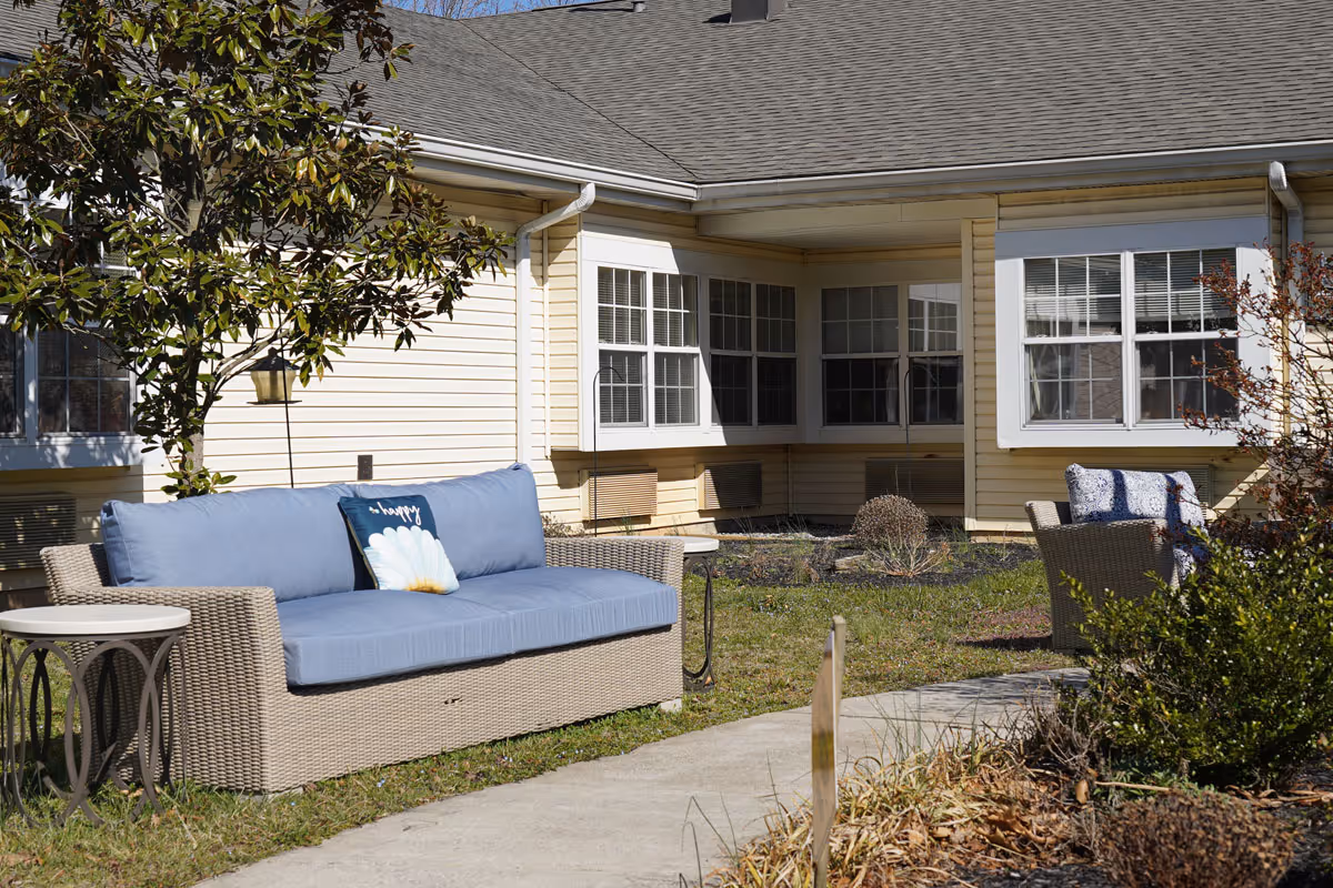 Outdoor patio area at a senior living facility with a wicker sofa featuring blue cushions and a decorative pillow, a small round side table, and a wicker chair with blue patterned cushions. The patio is surrounded by grass, bushes, and a tree, with the beige exterior of the building and multiple windows in the background.