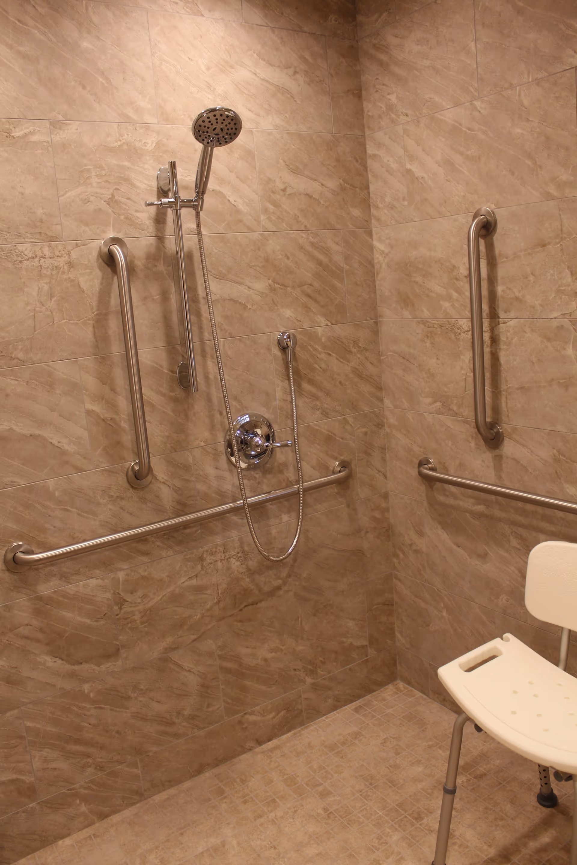 Accessible tiled shower with a handheld showerhead, multiple stainless steel grab bars, and a white shower chair.