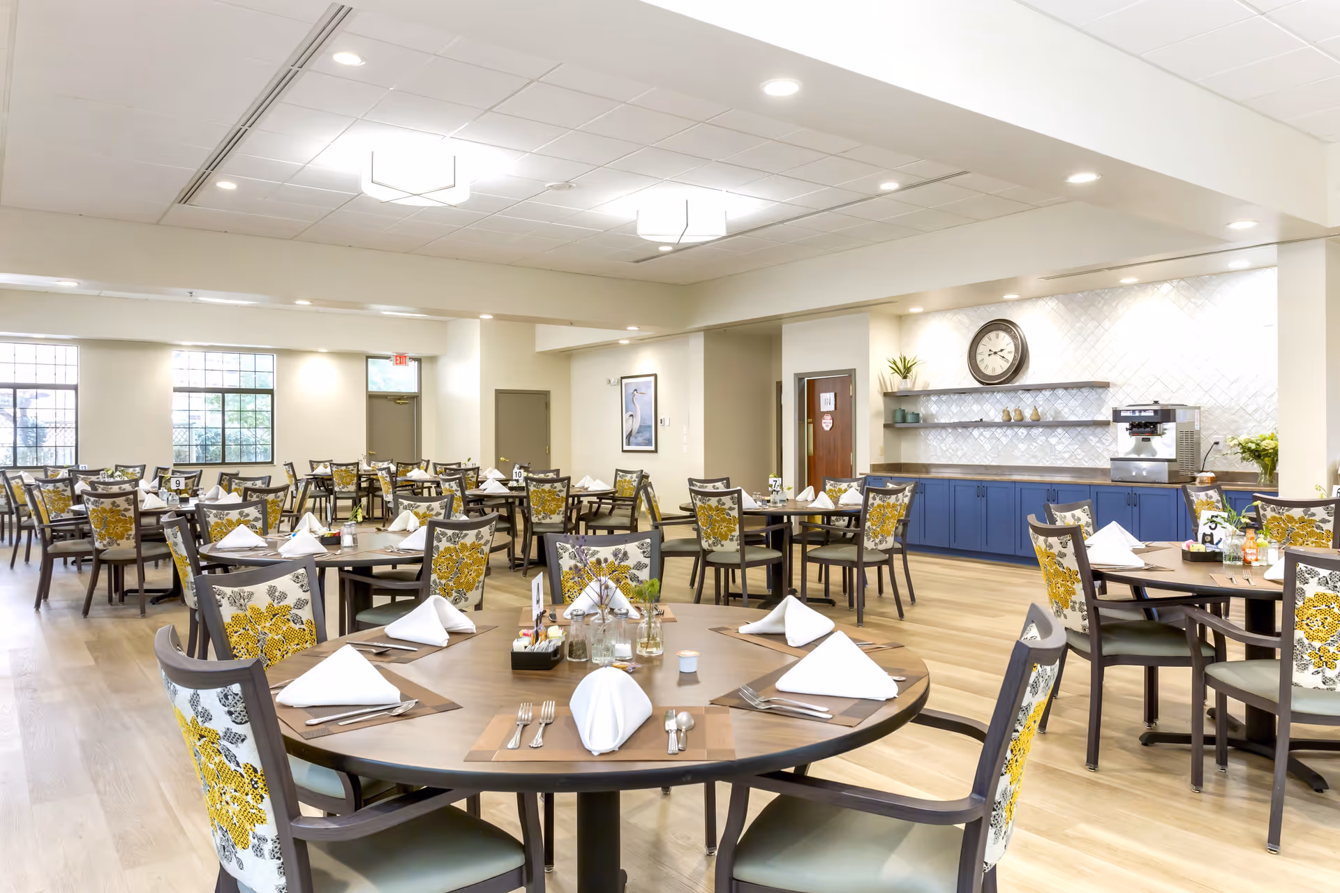 A bright and spacious dining room in The Montclair Senior Living facility featuring multiple round tables set with white folded napkins, silverware, and small decorative plants. Chairs have floral patterned upholstery in yellow and gray. The room has large windows, light wood flooring, and a wall with blue cabinets, shelves, a clock, and a coffee machine.