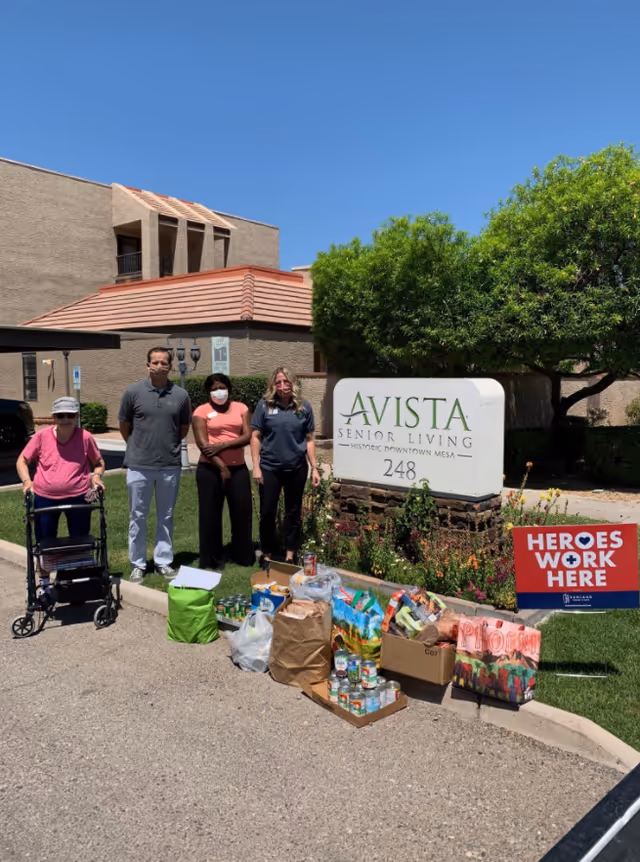 Four people stand outside the Avista Senior Living building beside its sign and a collection of donated food and care items.
