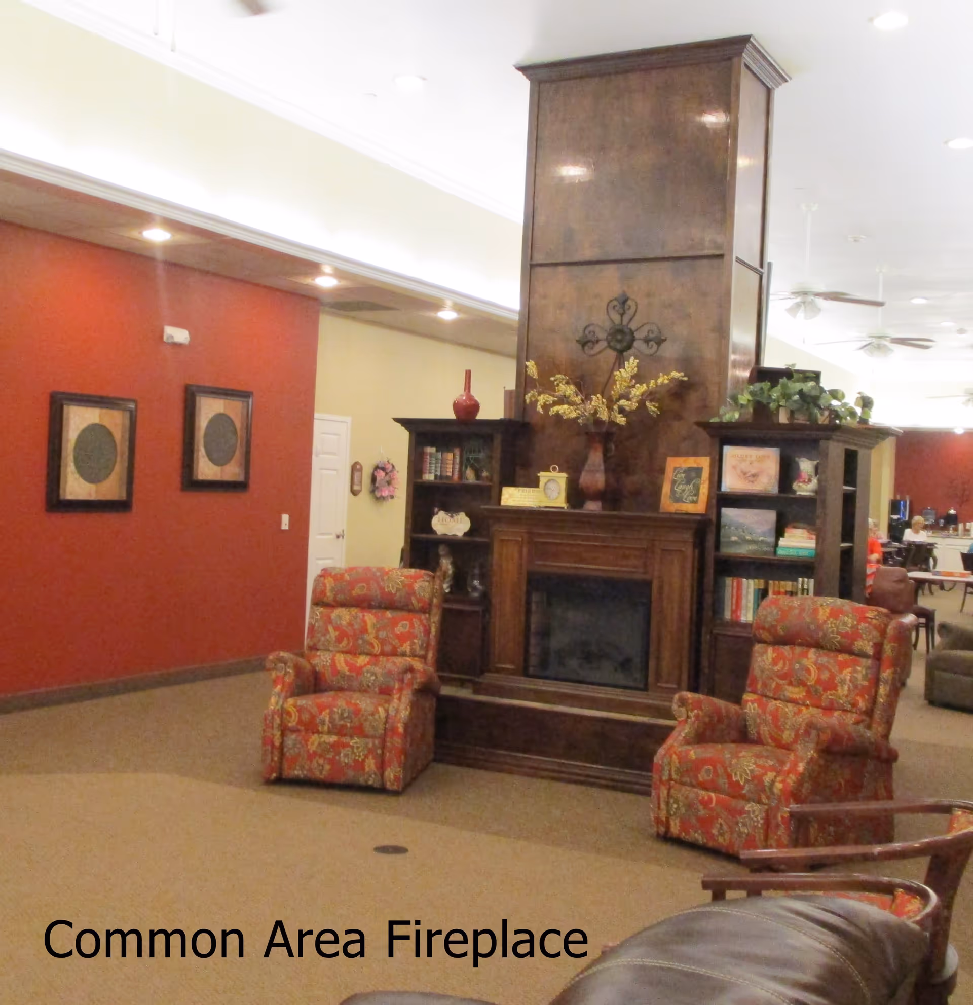 A cozy common area with a large wooden fireplace in the center, flanked by two patterned armchairs. The fireplace has built-in shelves on either side with decorative items and books. The walls are painted red and beige, with framed artwork on the red wall. Ceiling lights and fans are visible, and there are tables and chairs in the background.
