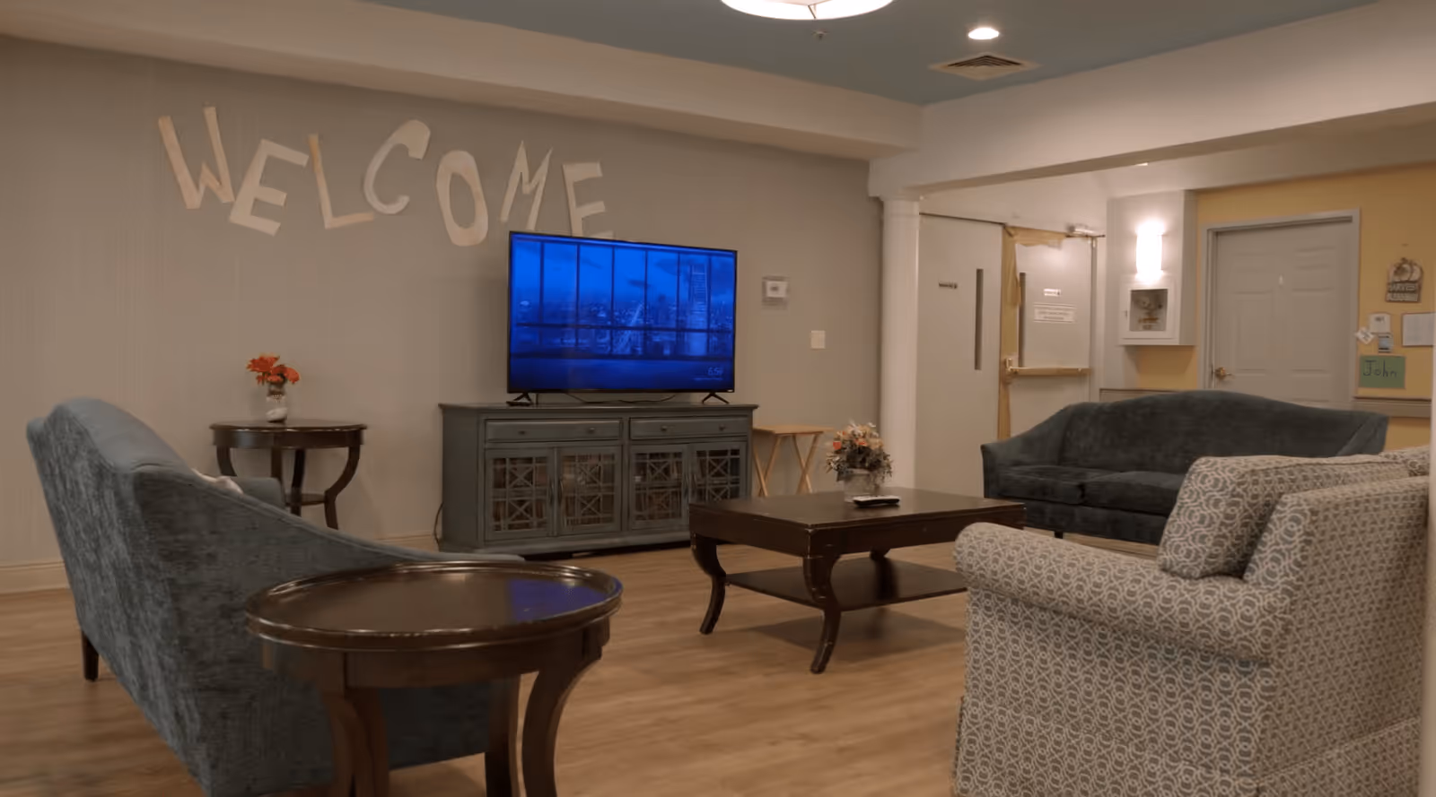 A cozy living room area in a senior living facility with a 'WELCOME' sign on the wall above a flat-screen TV on a cabinet. The room has a wooden coffee table, two blue upholstered sofas, a patterned armchair, and side tables with flower arrangements. The walls are light-colored with a door and some wall decorations in the background.