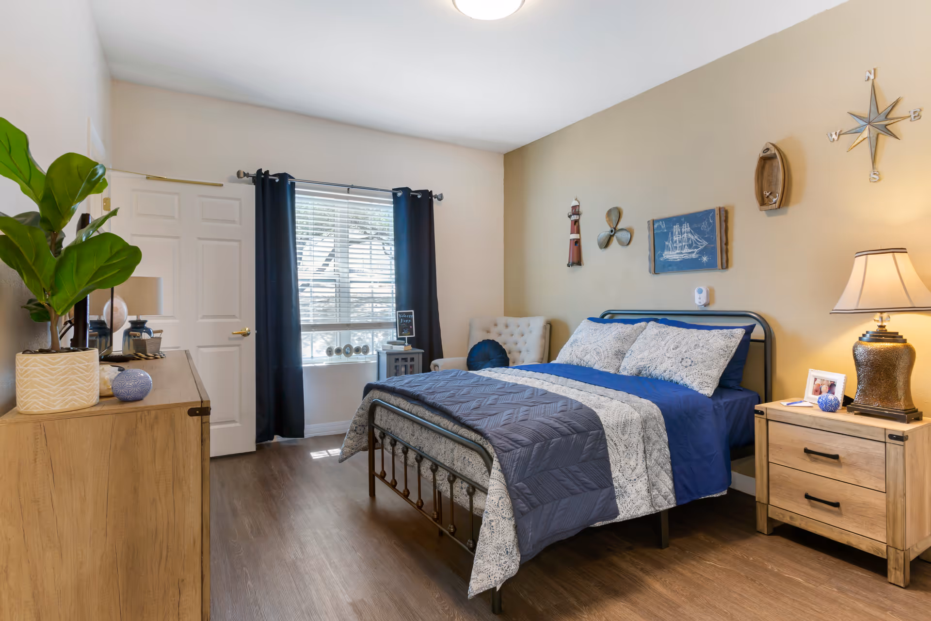 A cozy bedroom with a metal frame bed dressed in blue and white bedding. The room features a wooden dresser with a potted plant and decorative items, a nightstand with a lamp and framed photo, a window with dark curtains, and nautical-themed wall decorations including a compass rose, lighthouse, and ship artwork.