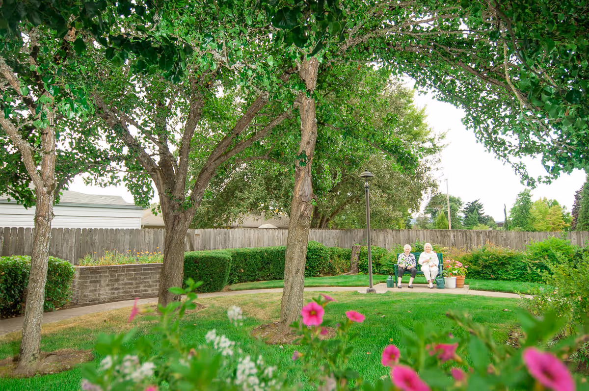 Two elderly women sitting on a bench in a lush garden area with green grass, trees, flowering plants, and a wooden fence in the background.