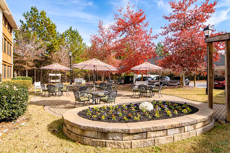 Outdoor patio area at Creekside Pines Retirement Community featuring round tables with chairs and striped umbrellas, surrounded by trees with red autumn leaves, a raised flower bed with colorful flowers, and a parking area with cars in the background.