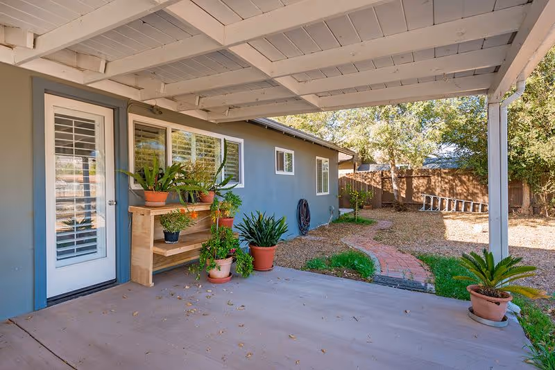Covered patio area with potted plants on a wooden shelf and on the ground, adjacent to a house with blue-gray walls and white-framed windows. A brick pathway leads through a backyard with trees and a wooden fence.