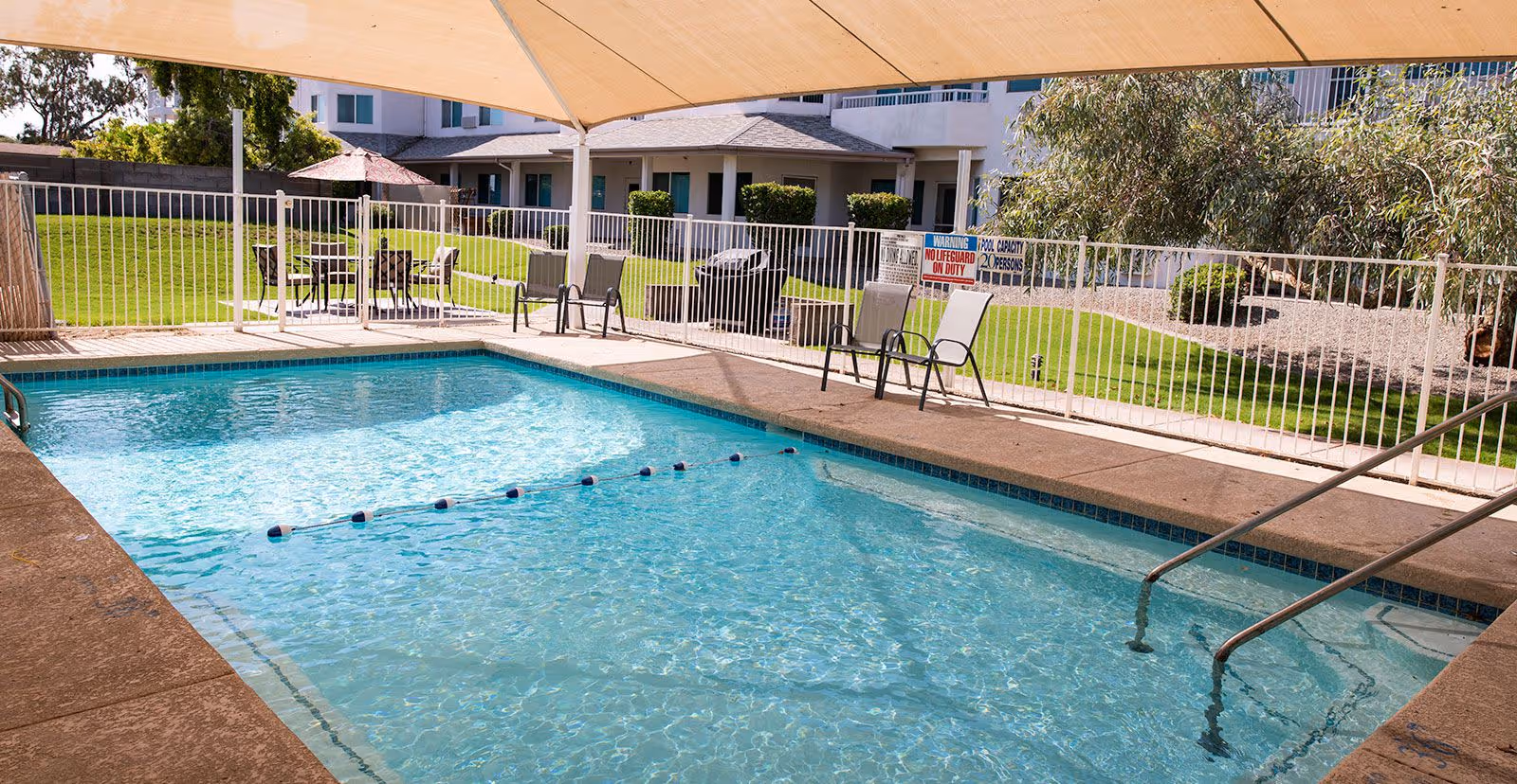 Outdoor swimming pool with clear blue water, surrounded by a concrete deck and white safety fence. Several chairs and tables with umbrellas are placed around the pool area. A large beige shade canopy covers part of the pool. In the background, there is a grassy area and a multi-story building with balconies and windows.