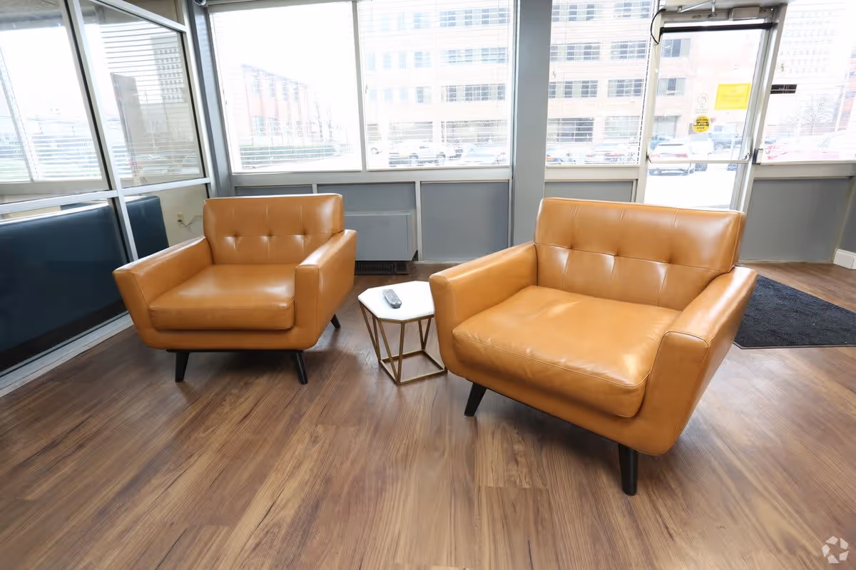 A bright interior room with two tan leather armchairs positioned around a small white hexagonal side table with a remote control on it. The room has large windows with a view of buildings and a parking lot outside. The floor is wood, and there is a black mat near a glass door.