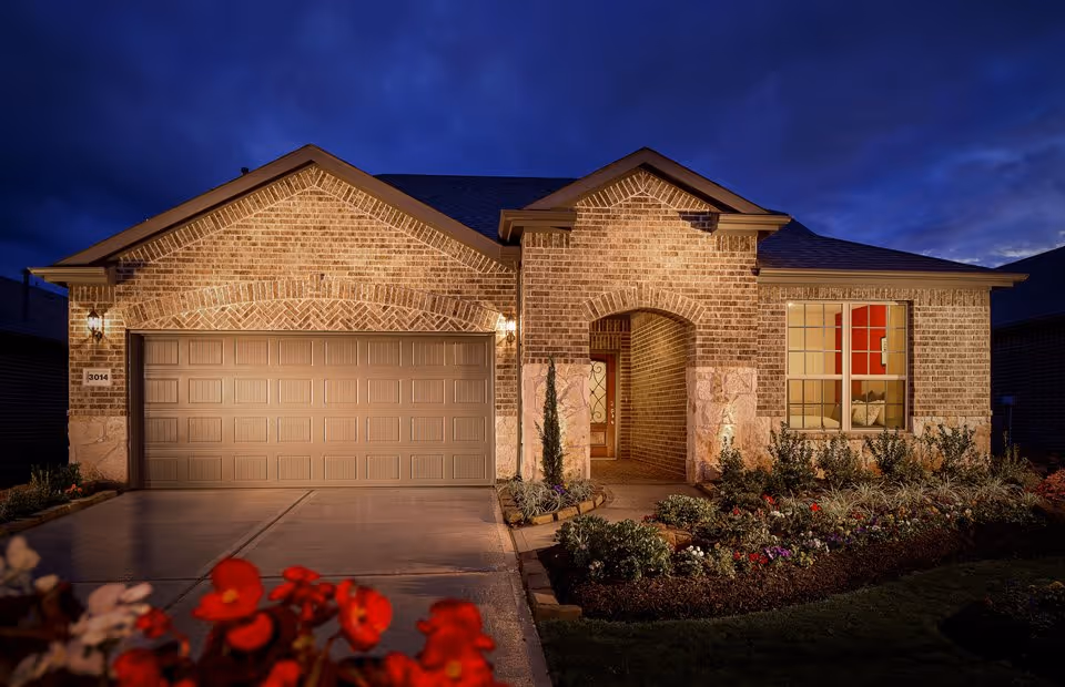 A single-story brick house at dusk with a two-car garage, illuminated exterior lights, a front porch with an arched entryway, and a well-maintained garden with flowers in the foreground.