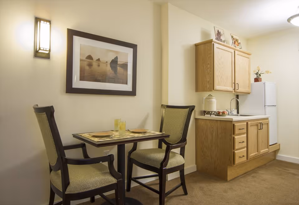 Small dining area with a table set for two, featuring two upholstered chairs. Adjacent to the dining area is a compact kitchen with wooden cabinets, a countertop with a sink, a small stove, and a white refrigerator. A framed picture hangs on the wall above the dining table, and a wall-mounted light fixture provides illumination.