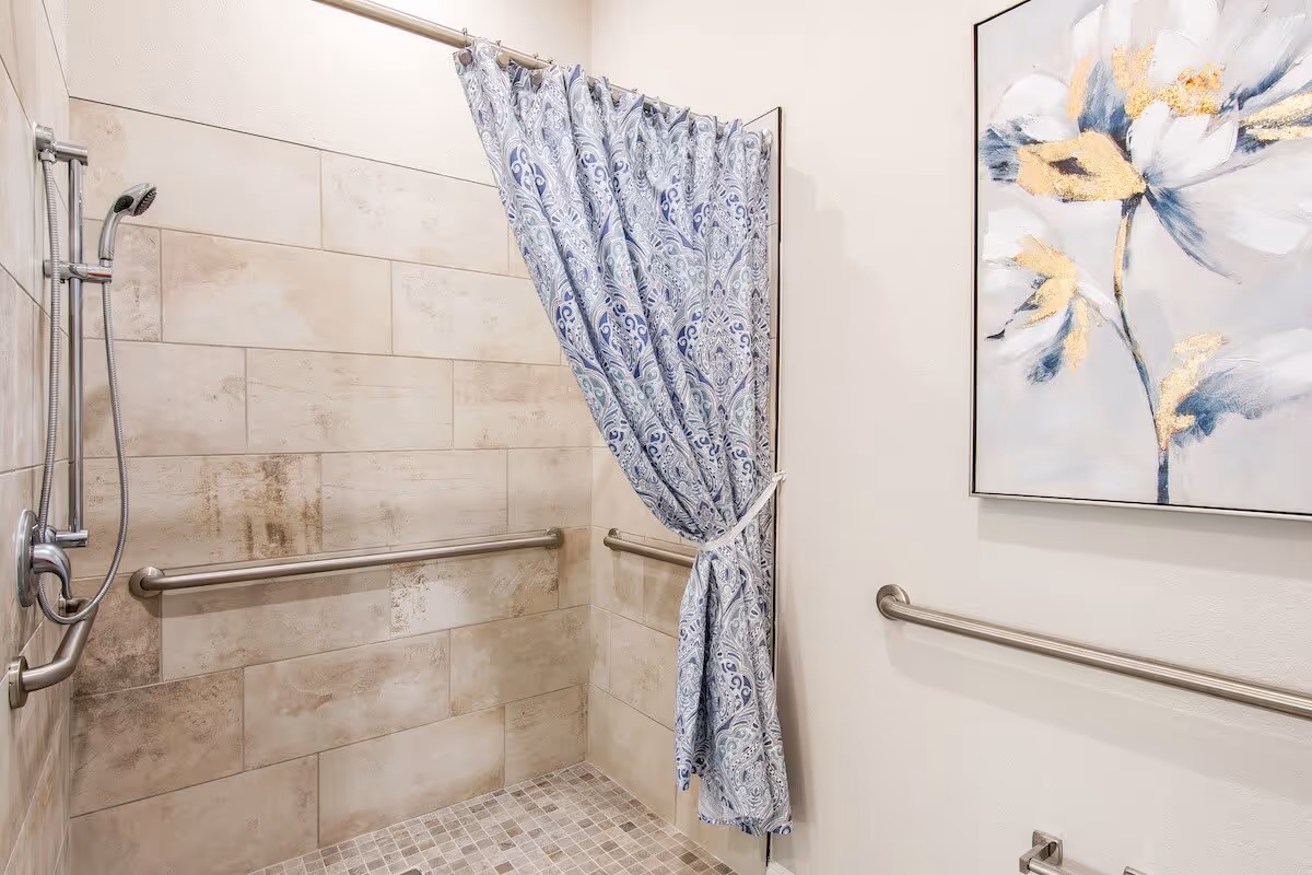 Accessible tiled walk-in shower with grab bars, a handheld showerhead, and a patterned curtain next to floral wall art.