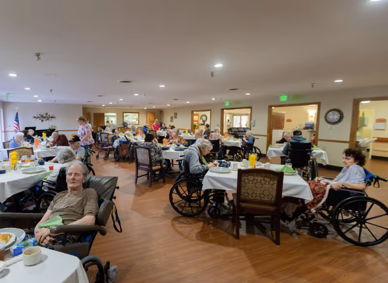 Large dining room with many elderly residents seated at tables, several in wheelchairs, and staff serving meals.