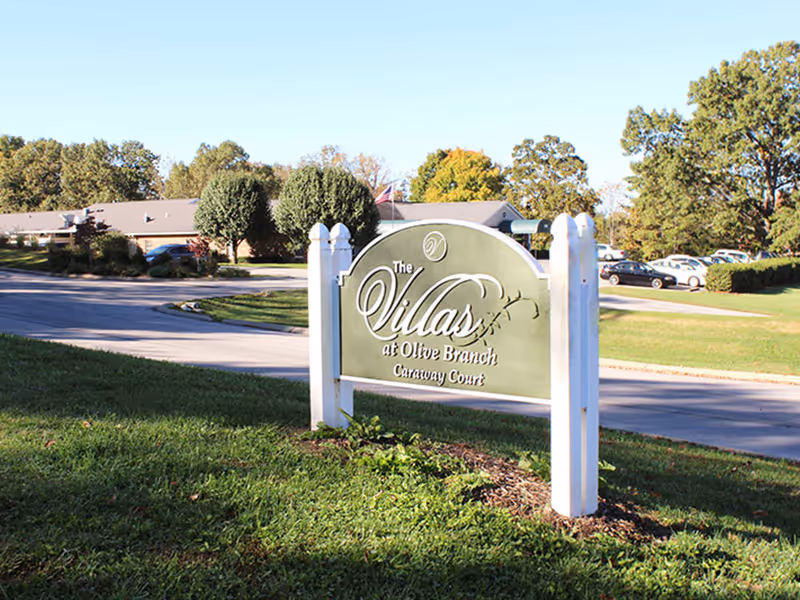 Outdoor view of a senior living facility entrance sign that reads 'The Villas at Olive Branch Caraway Court' with a driveway, parked cars, trees, and a building in the background under a clear sky.