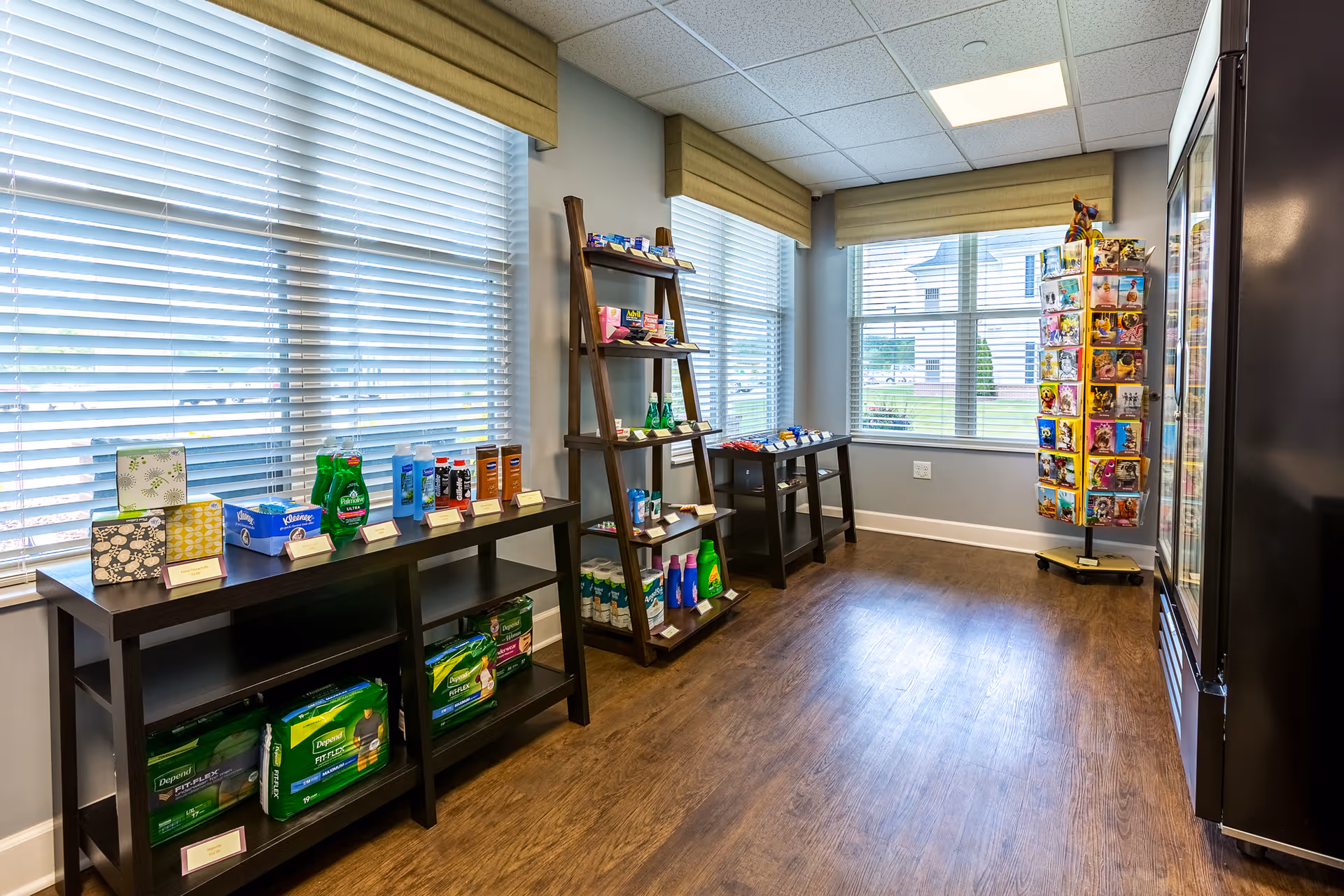 A small retail area inside a senior living facility with shelves and racks displaying various personal care products, snacks, and greeting cards. The room has large windows with blinds, wood flooring, and bright overhead lighting.