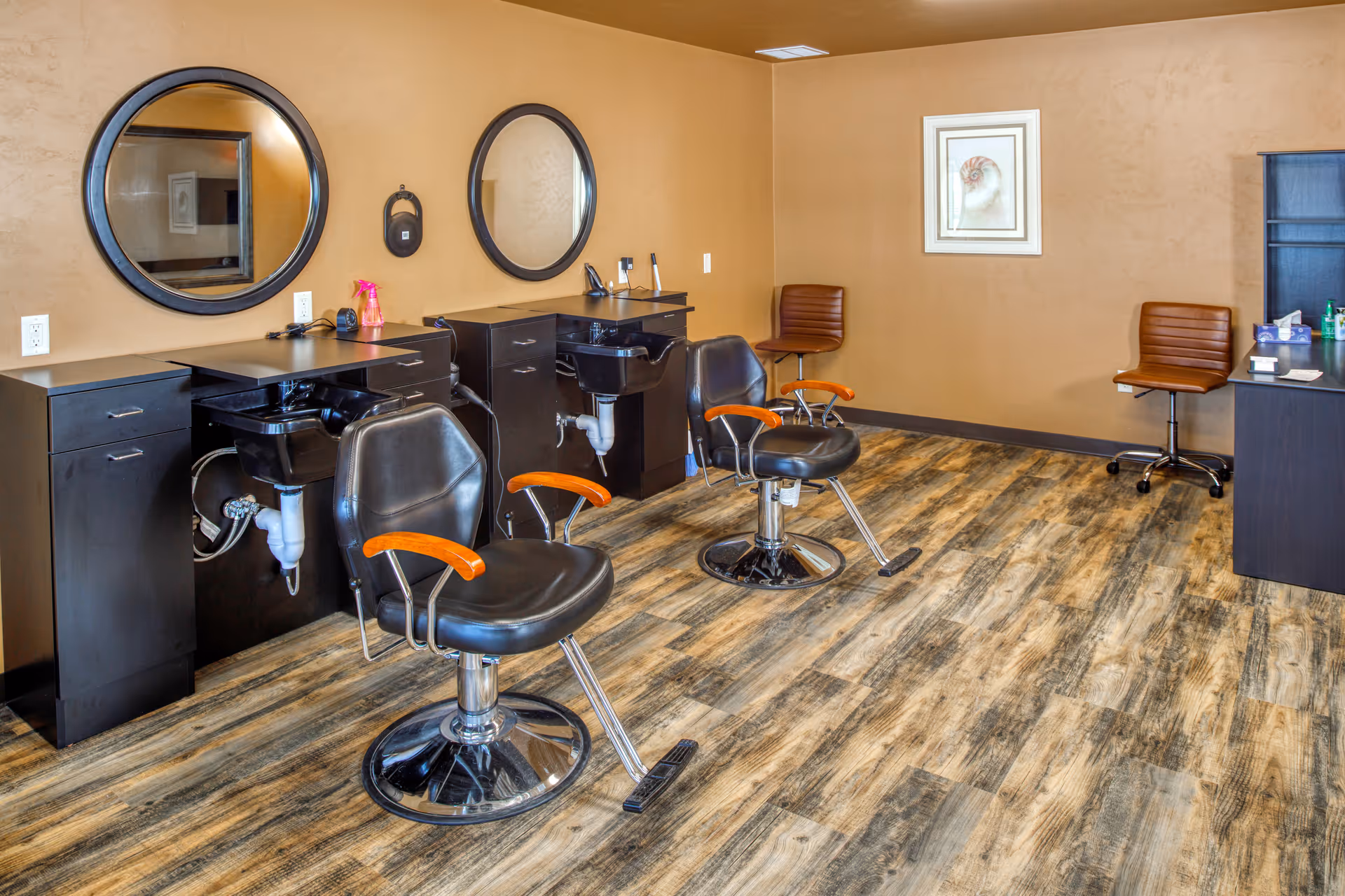 Interior of a salon area with two black salon chairs with wooden armrests in front of two black washbasins and round mirrors mounted on a beige wall. The floor has a wood-like pattern, and there are two brown chairs and a black cabinet with various items on the right side of the room.