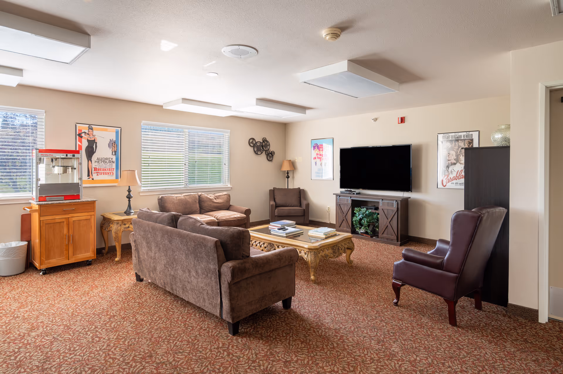 Bright communal living room with sofas, armchairs, a coffee table, television and a popcorn machine near windows.