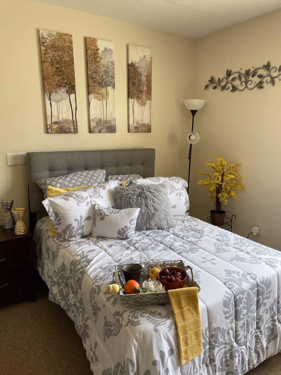 A neatly made bed with a gray tufted headboard and white and gray patterned bedding. The bed has multiple pillows including a fluffy gray one and a small decorative pillow. A tray with a cup, a glass, fruit, and a yellow cloth is placed on the bed. On the left side, there is a dark wooden nightstand with two decorative vases. On the wall above the bed, there are three vertical paintings of trees in autumn colors. To the right, there is a tall floor lamp and a potted plant with yellow flowers. The walls are painted beige.