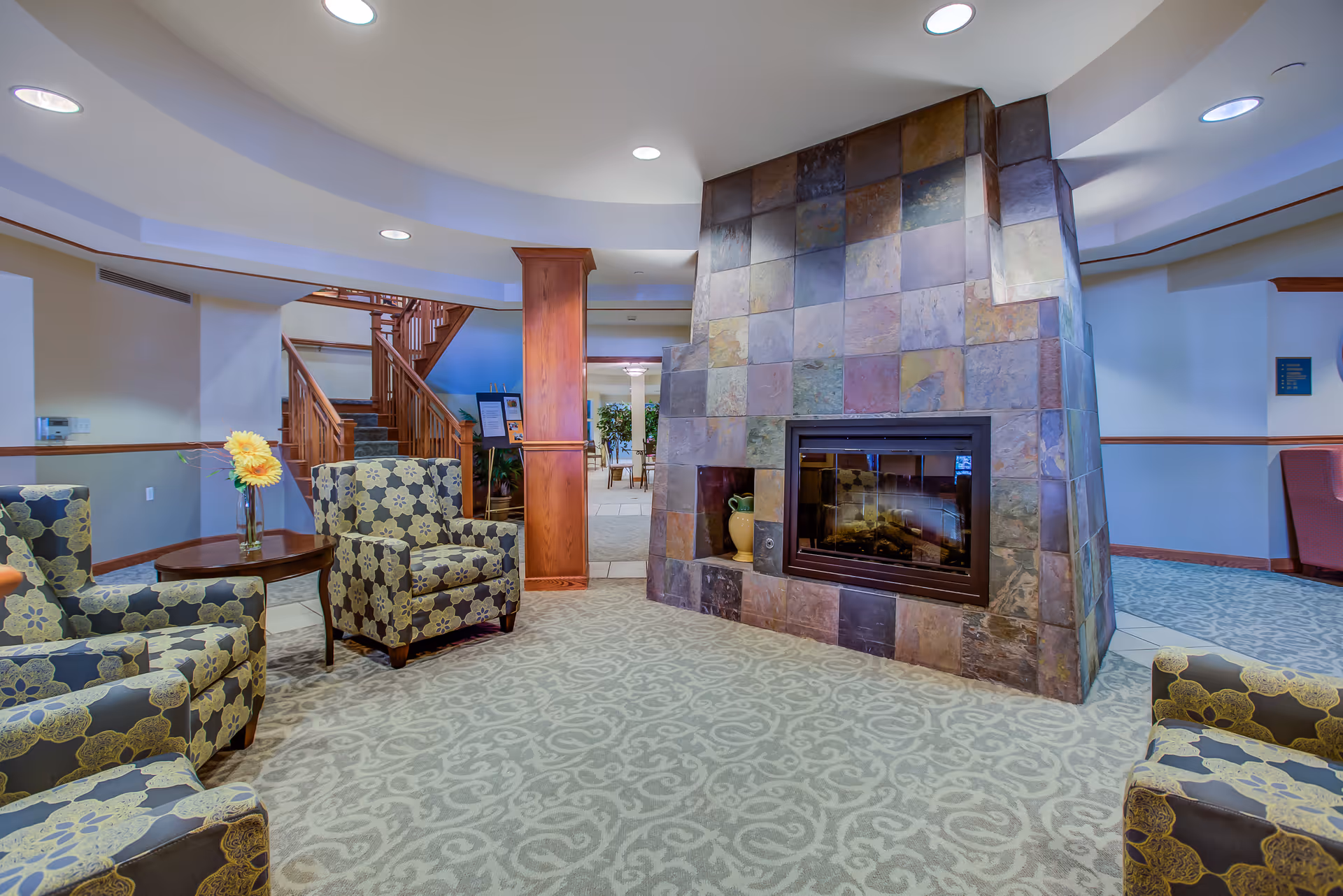 A cozy living room area in Town Village Sterling Heights featuring patterned armchairs arranged around a tiled fireplace. The room has a carpeted floor with a decorative pattern, a wooden staircase in the background, and a small table with a vase of yellow flowers.
