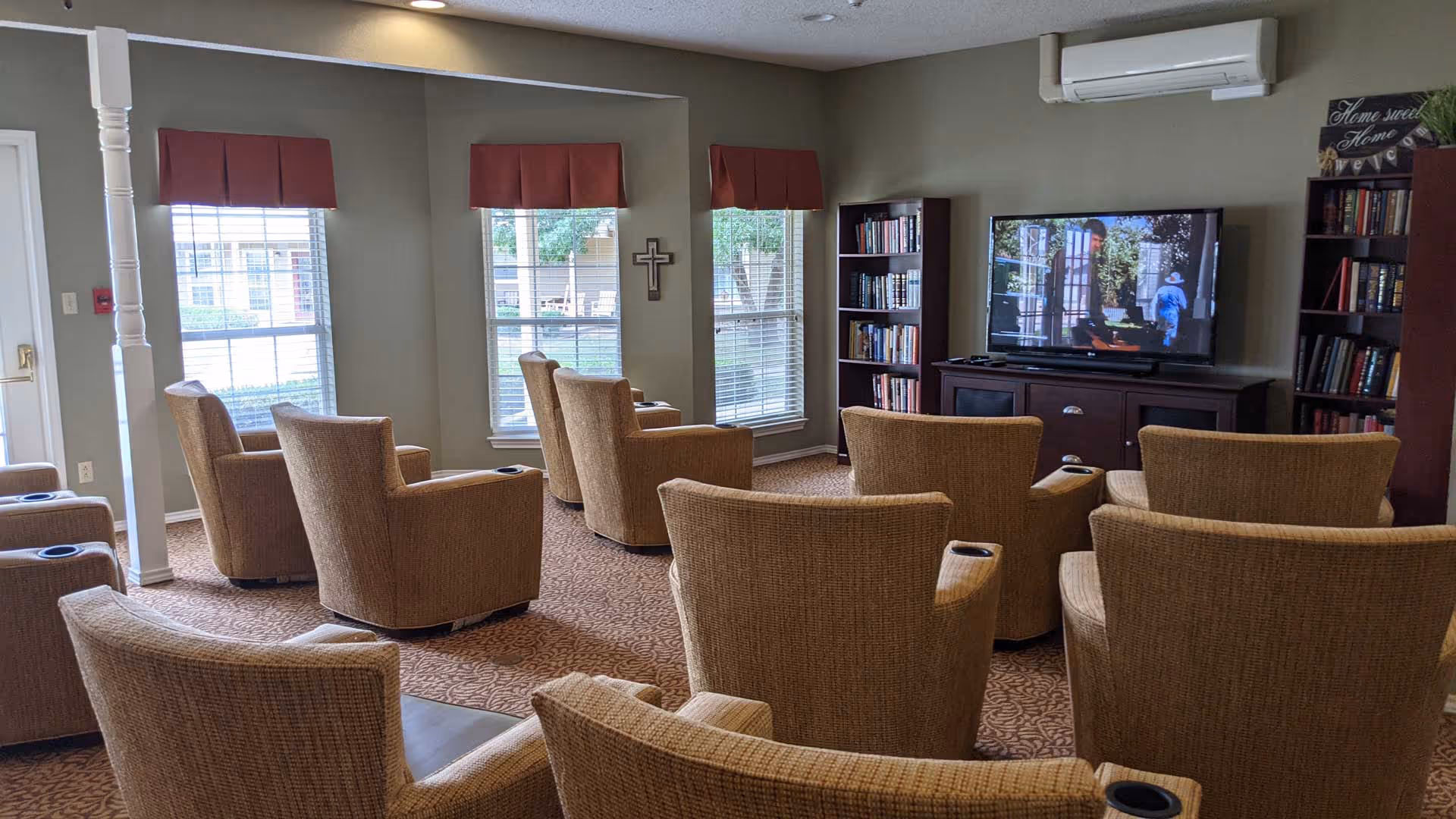 A senior living common room with multiple beige armchairs arranged facing a large TV and bookshelves by windows.