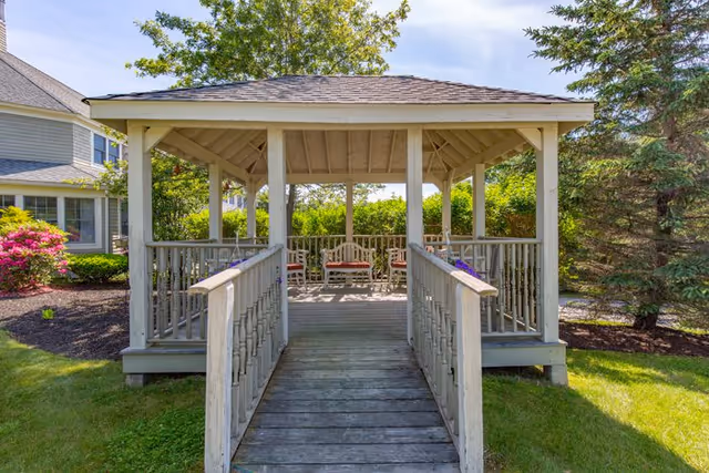A wooden gazebo with a ramp leading up to it, surrounded by green grass, trees, and flowering bushes. The gazebo has a shingled roof and contains several chairs inside. The sky is clear and blue.