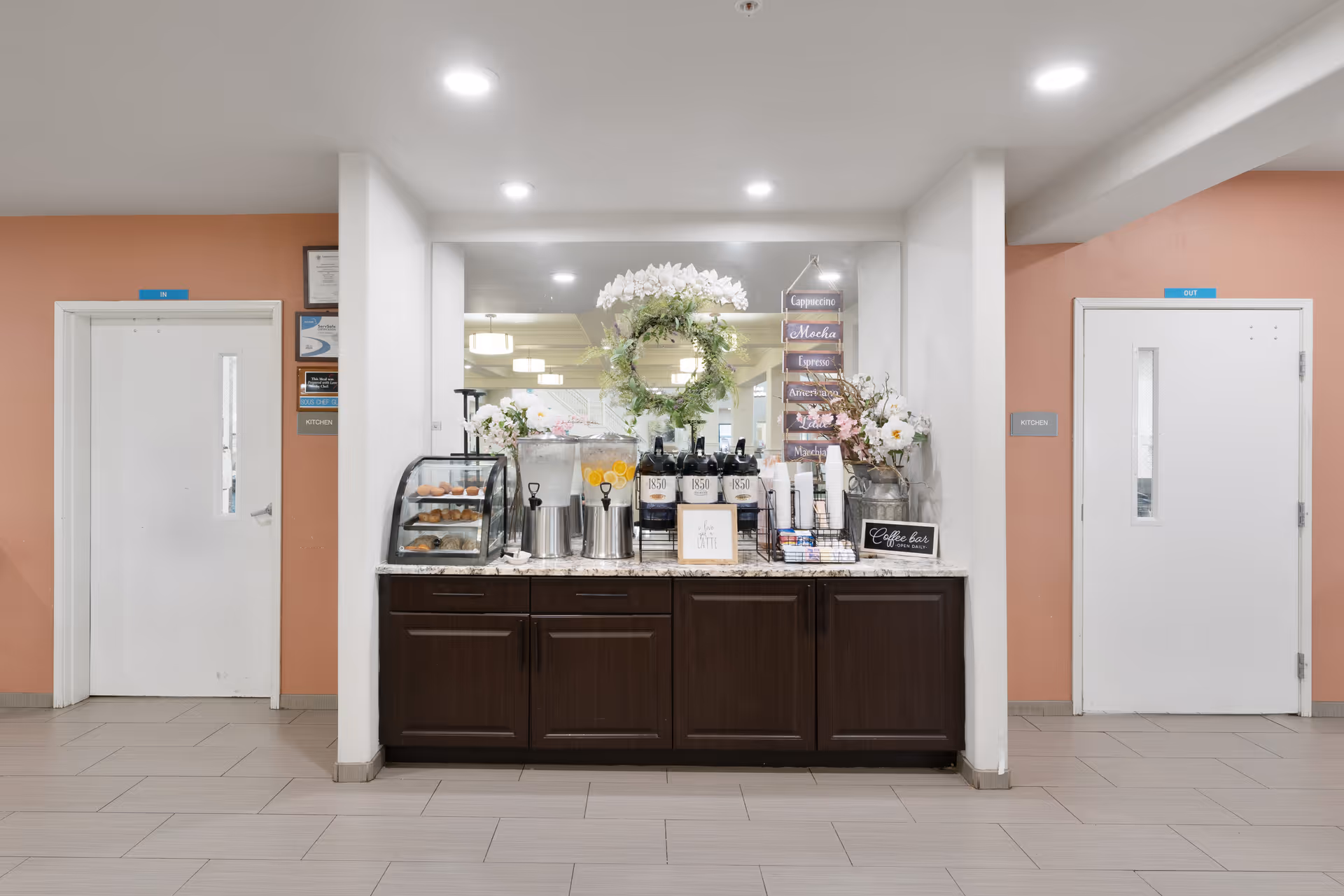 A coffee bar setup in a hallway with two white doors on either side labeled 'Kitchen' and 'Out'. The coffee bar features a display case with pastries, beverage dispensers, coffee cups, and decorative flowers with a large mirror behind it. The walls are painted peach and white, and the floor is tiled.