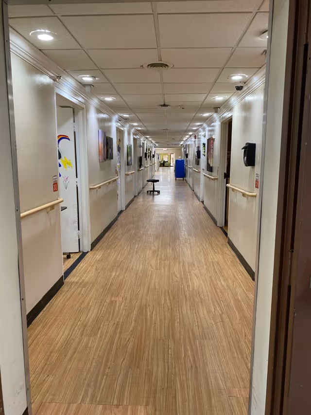 A long, well-lit hallway in a nursing center with light-colored walls and wood-patterned flooring. The hallway has handrails on both sides and several doors leading to rooms. There are paintings and decorations on the walls, a black stool in the middle of the hallway, and a blue cart further down the corridor.