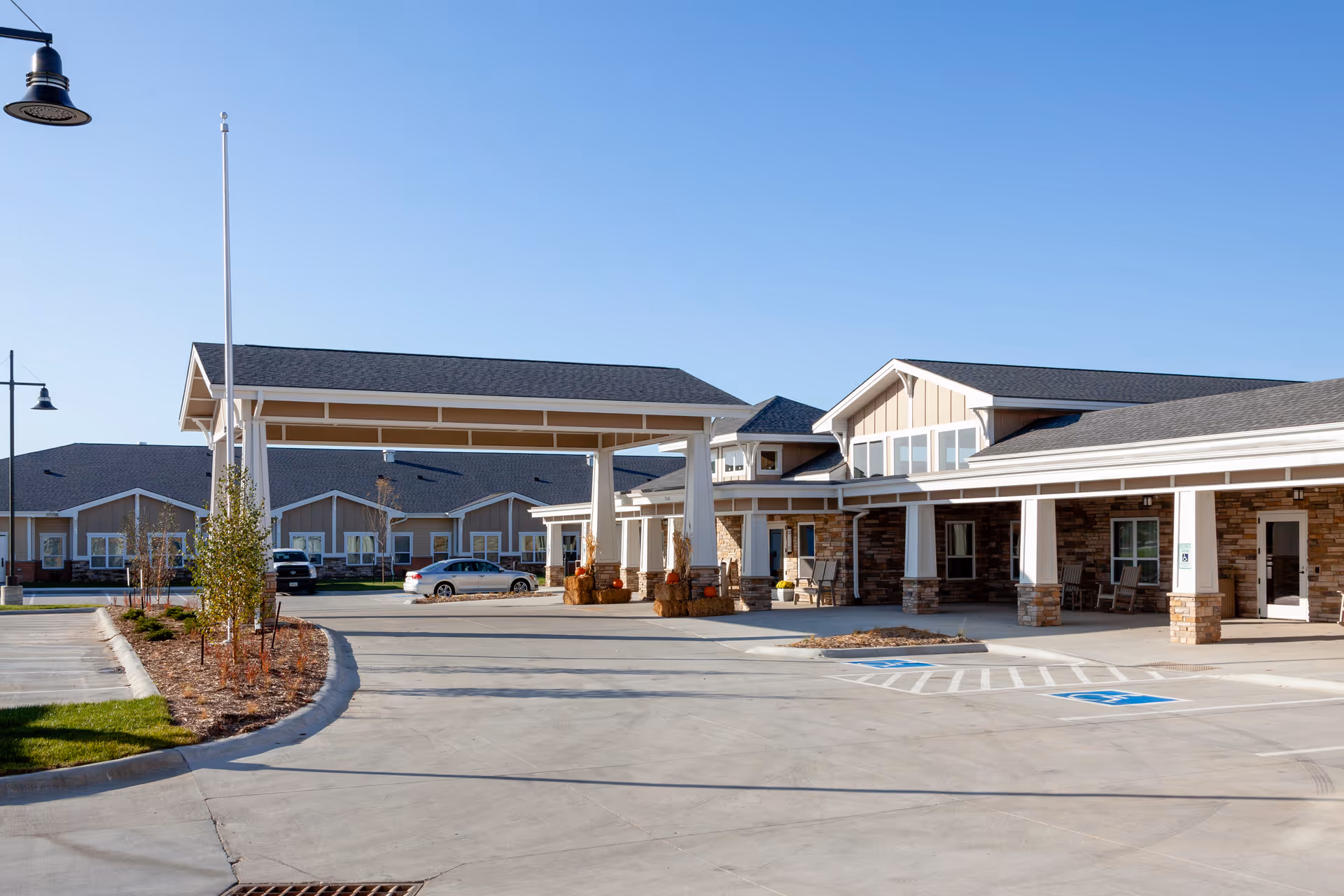 Exterior view of Cedarhurst Senior Living of La Vista showing a large covered entrance with a driveway and parking spaces, including handicapped spots. The building features a combination of stone and siding with multiple windows and a clear blue sky above.