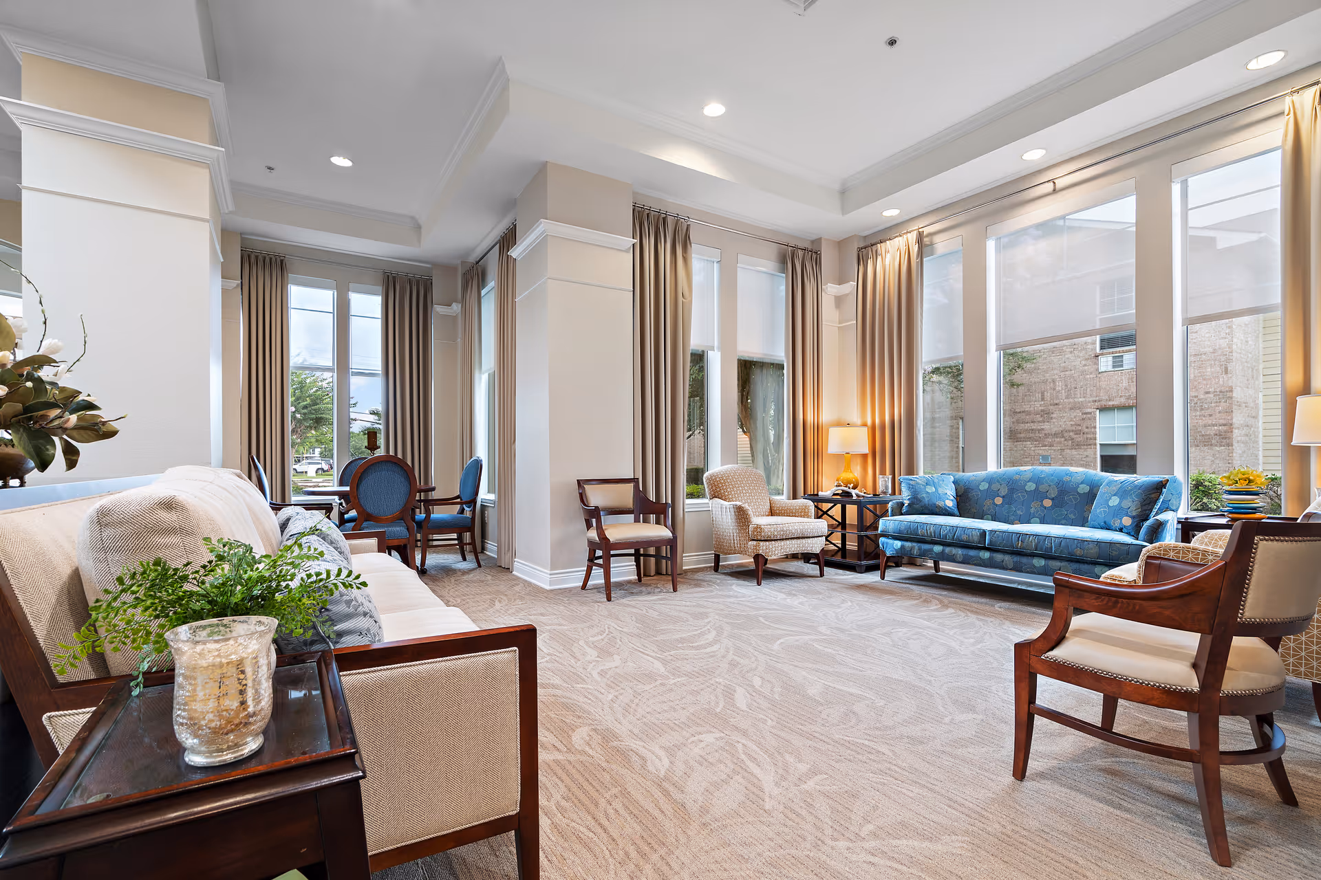 Bright, airy common lounge with upholstered sofas and armchairs, large windows, side tables and neutral carpeting.