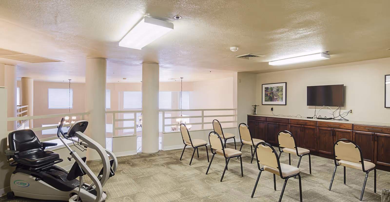 A spacious room with beige walls and carpeted floor featuring a row of chairs facing a wall-mounted flat screen TV above a long wooden cabinet. To the left, there is exercise equipment including a recumbent bike. The room has large pillars and overlooks a lower level with windows and hanging light fixtures.