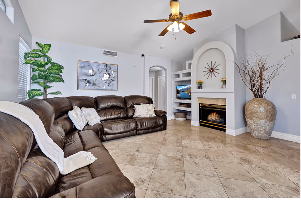 A spacious living room with a large brown leather sectional sofa adorned with white and patterned pillows and a white throw blanket. The room features a ceiling fan with lights, a tiled floor, a modern fireplace with a decorative clock above it, built-in shelves with a TV and decorative items, a large potted plant near the window, and a large vase with tall branches in the corner.