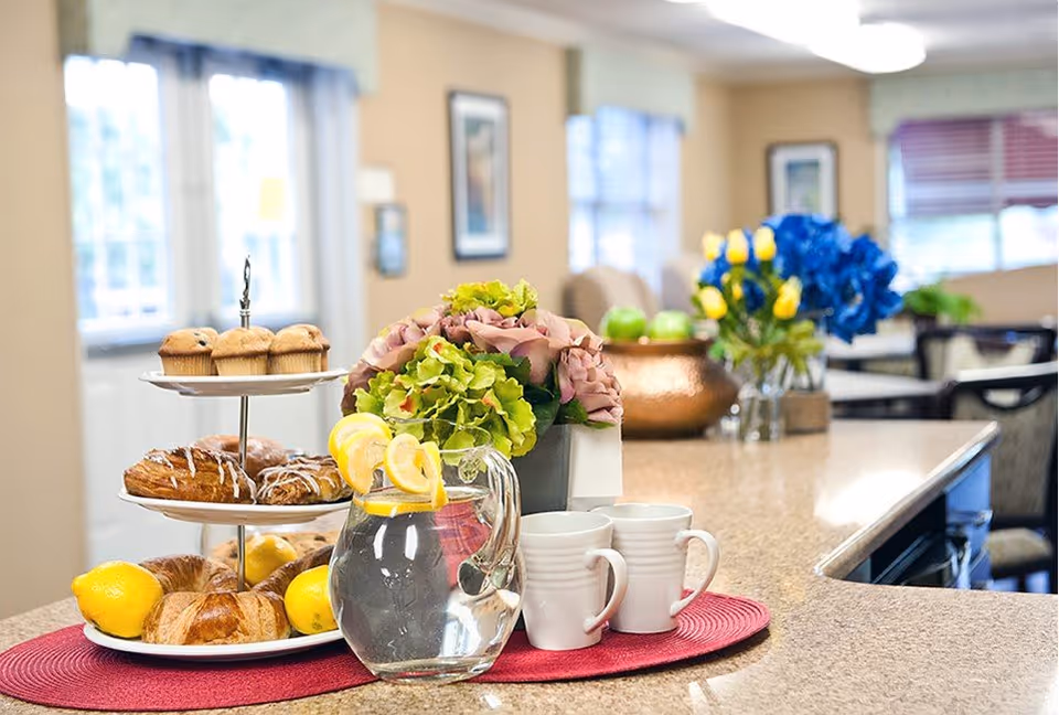 A kitchen or dining area countertop with a three-tiered tray holding muffins, pastries, and croissants surrounded by lemons. A glass pitcher of water with lemon slices and two white mugs sit on a red placemat. In the background, there are flower arrangements and a dining area with chairs and tables.