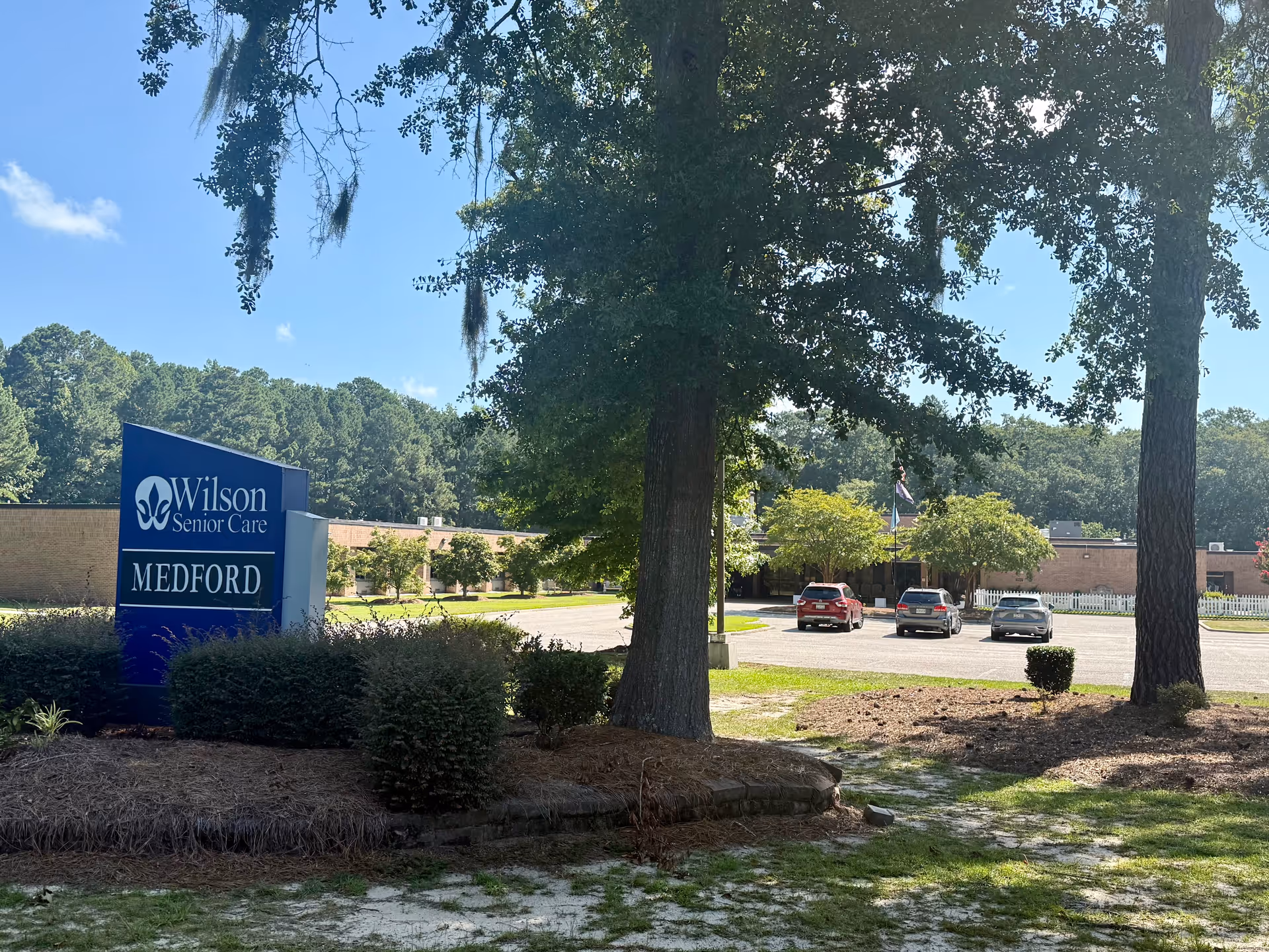 Exterior view of Wilson Senior Care Medford building with a parking lot and several cars, surrounded by trees and greenery under a clear blue sky.