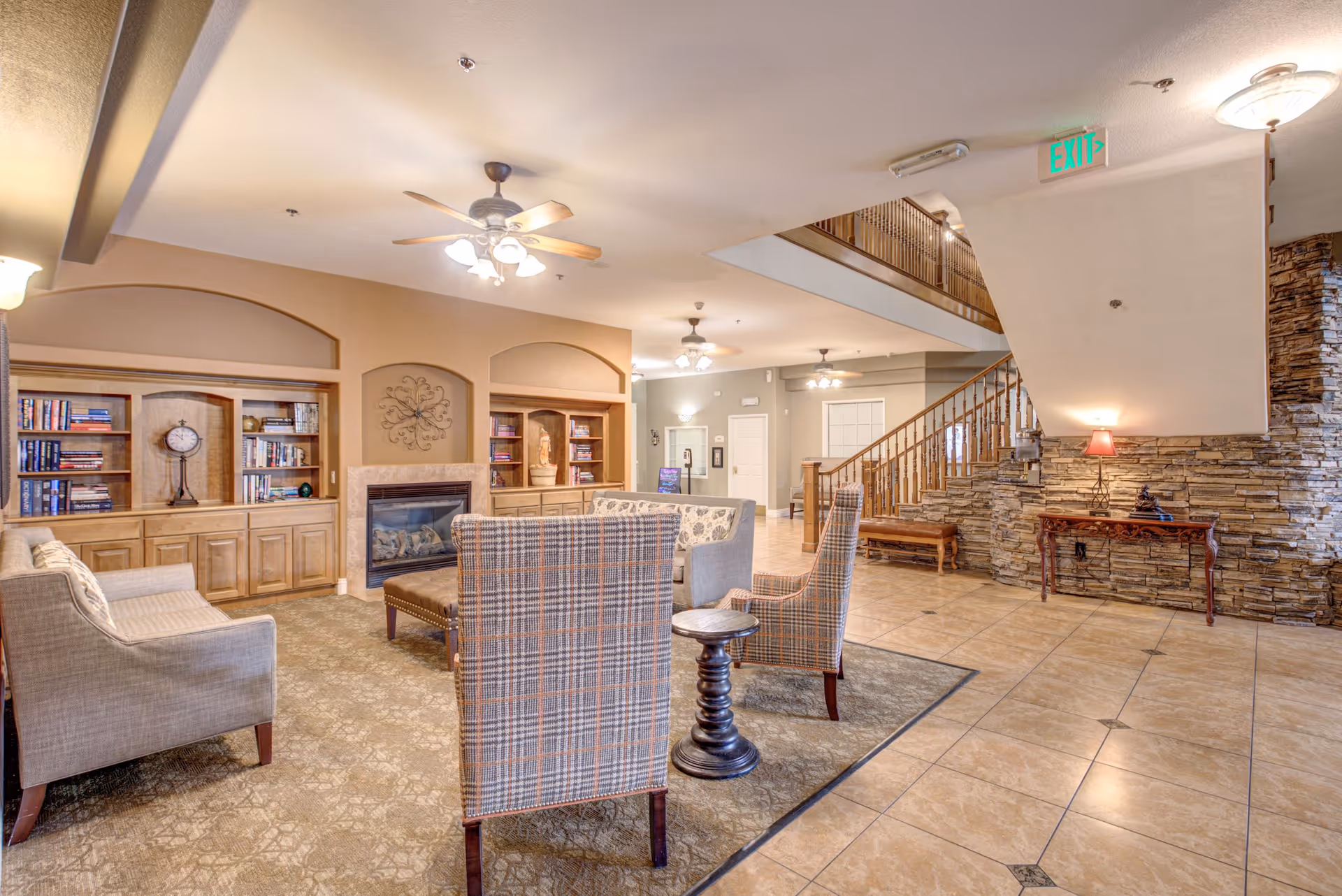 A spacious lounge with sofas and armchairs arranged around a fireplace and built-in bookshelves, with a staircase and tiled floor in the background.