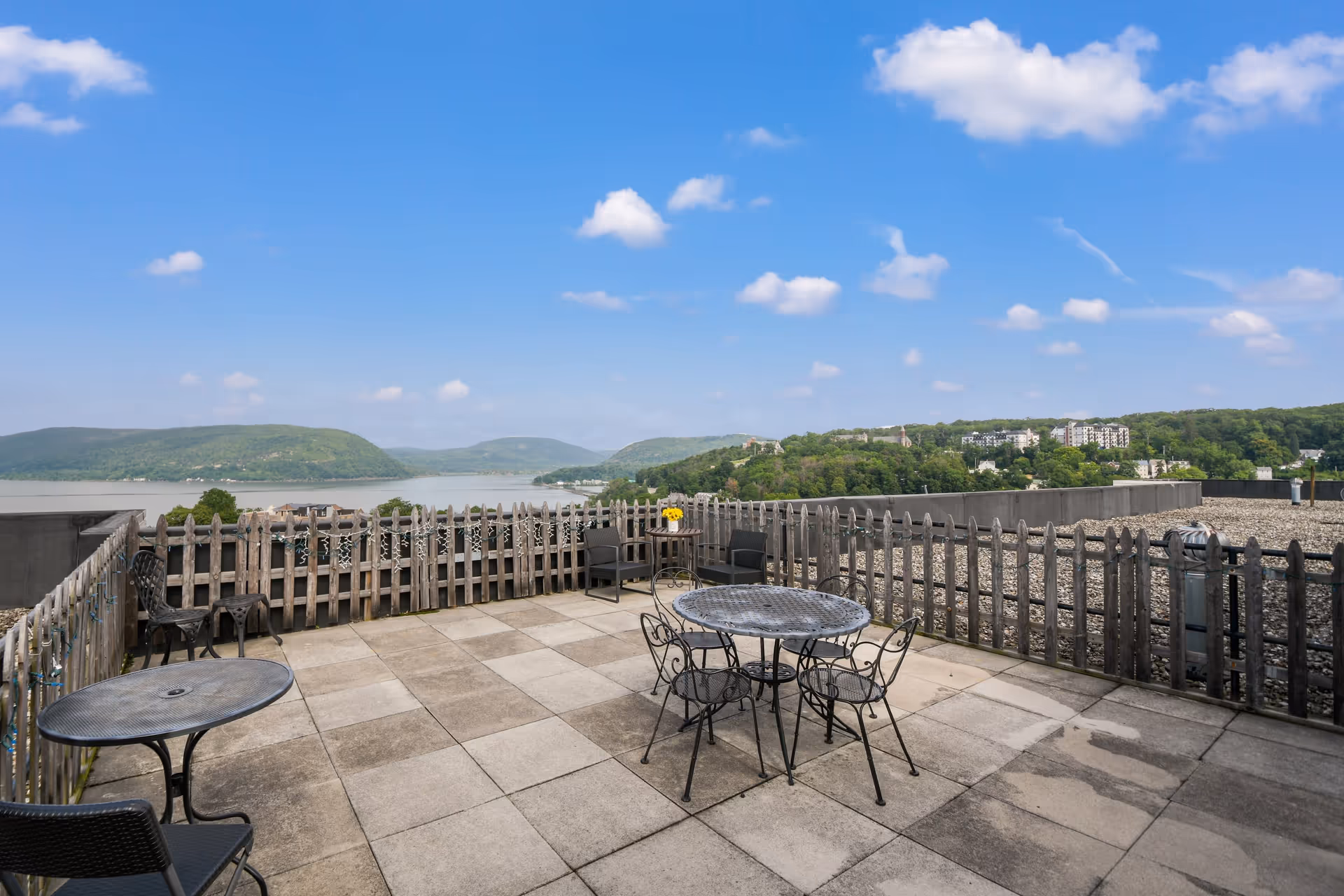 Outdoor patio area with metal tables and chairs on a tiled floor, surrounded by a wooden picket fence. The patio overlooks a scenic view of a river, green hills, and a partly cloudy blue sky.