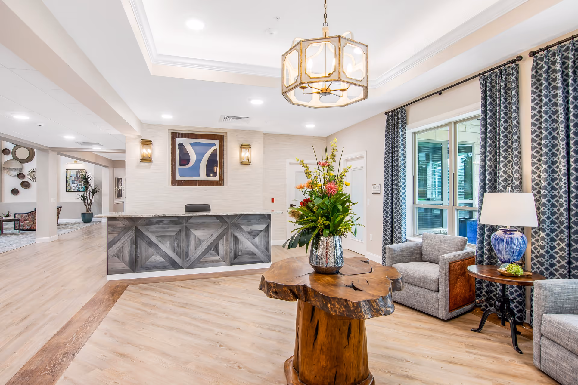A bright and welcoming senior living facility reception area with a wooden front desk, a large wooden table with a floral arrangement, two gray armchairs, a side table with a blue lamp, patterned curtains, and modern ceiling lights.