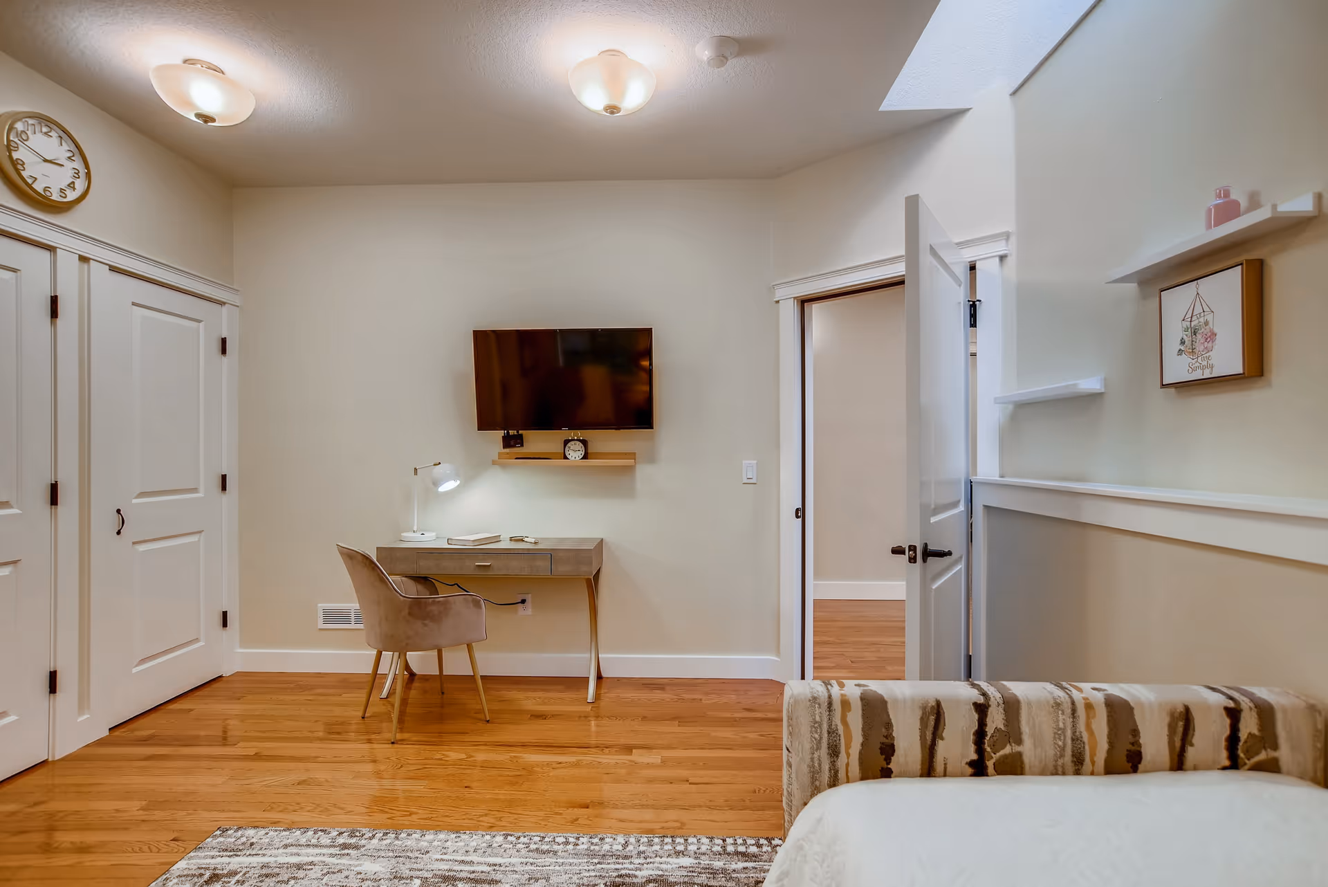 A cozy room with wooden flooring featuring a small desk with a chair and a desk lamp, a wall-mounted TV above a small shelf with a clock, a partially visible bed with a patterned blanket, a wall clock, and an open door leading to another room. The walls are light-colored, and there are two ceiling lights providing illumination.