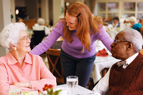 A caregiver interacts with two elderly residents at a dining table in a senior living facility.